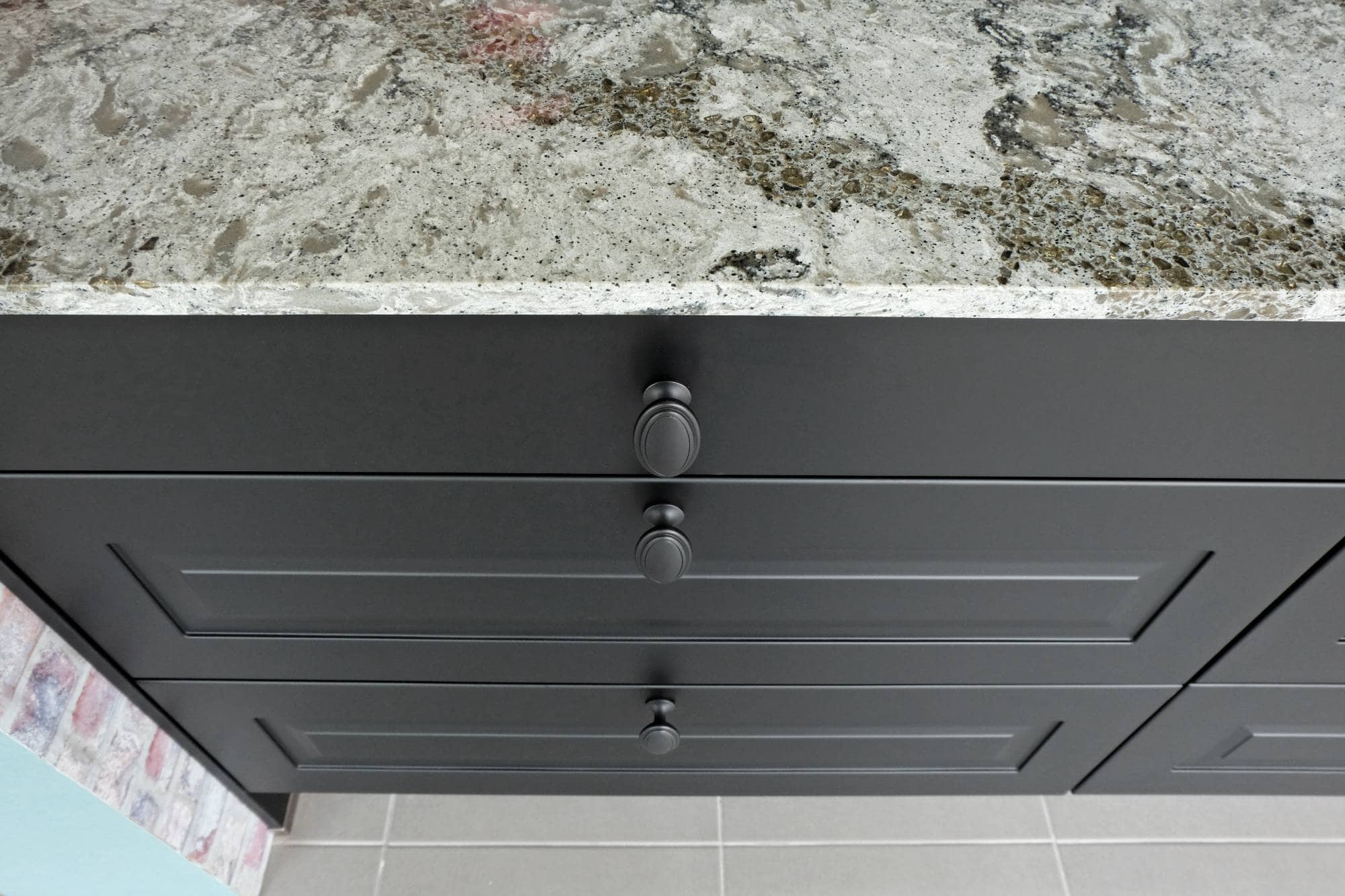 Close-up view of a modern kitchen countertop with a speckled gray and white stone surface above three matte black drawers, each with a round metal knob. The floor is light gray tile—perfect for homes in MI or after painting services Macomb & Oakland Counties, MI.