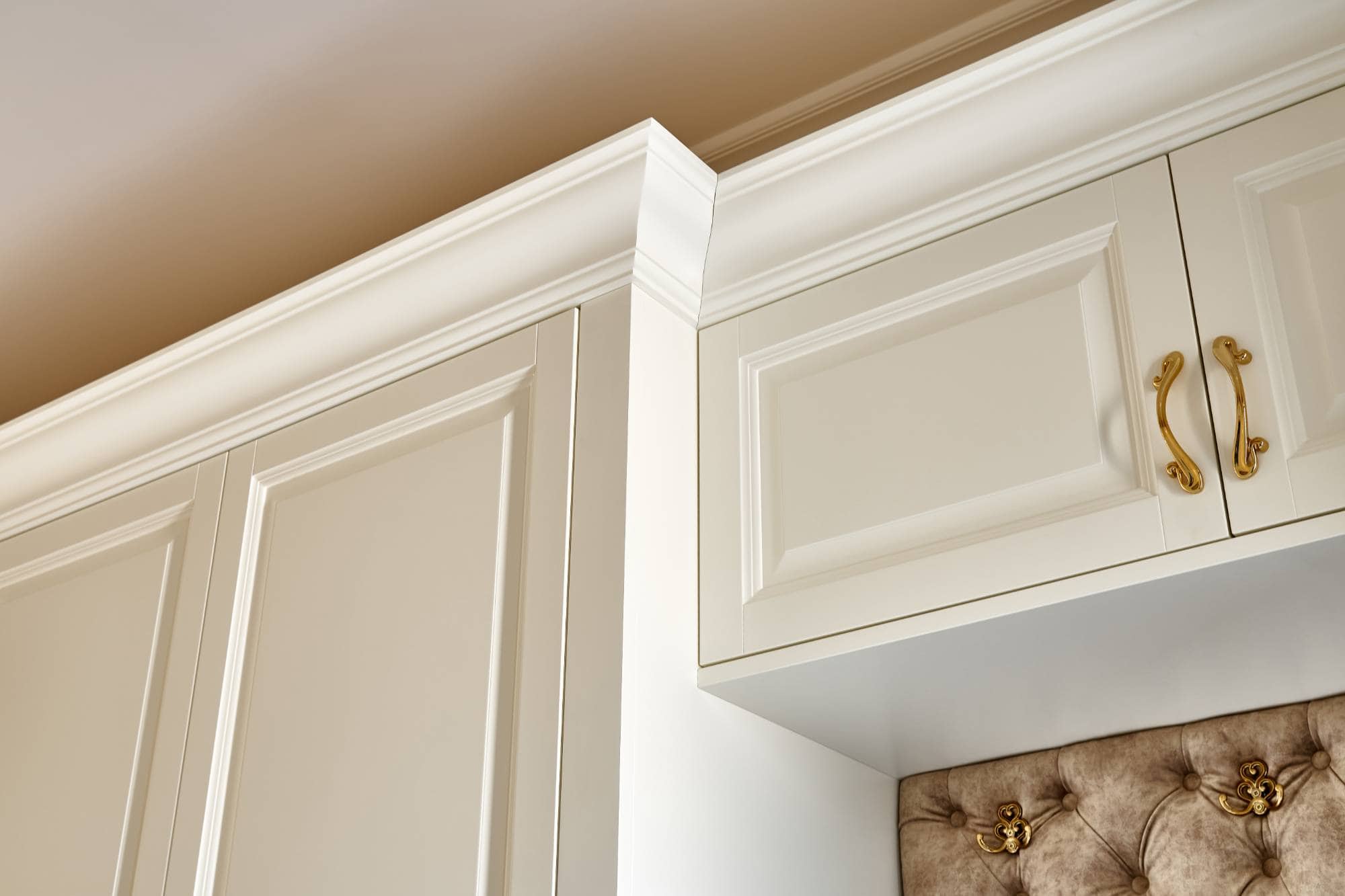 Close-up of elegant white kitchen cabinets with gold handles and crown molding against a beige ceiling, featuring detailed paneling. Perfect inspiration for those seeking painting services Macomb & Oakland Counties for classic design transformations.
