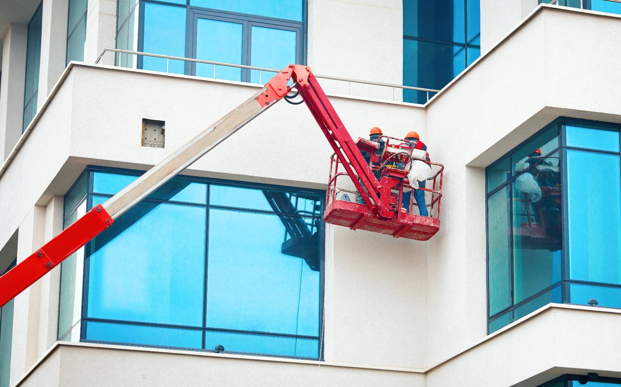 Two construction workers in safety helmets stand on a red lift platform, providing painting services Macomb & Oakland Counties as they work on the exterior windows of a modern building with large blue-tinted glass panels.
