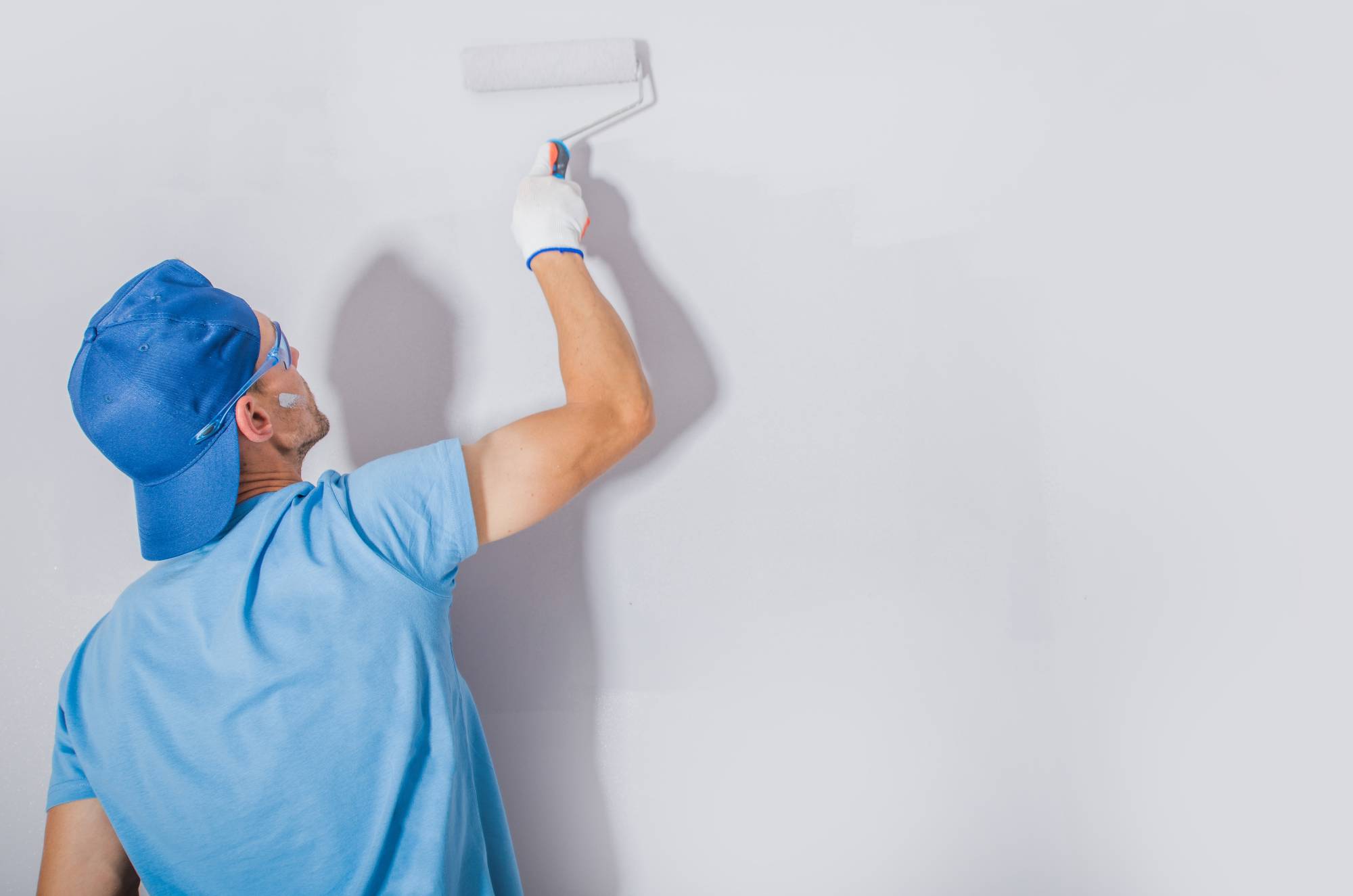 A Painter Macomb & Oakland Counties, MI, wearing a blue cap, blue shirt, and white gloves paints a white wall with a roller. The individual is facing the wall, with only their back and arm visible.