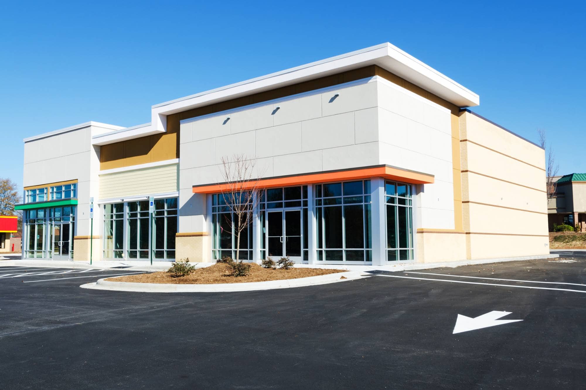 A modern, empty commercial building with large glass windows and doors stands under a bright blue sky in MI, its spacious parking lot ready for customers—ideal for any business, including a Painter Macomb & Oakland Counties,MI.