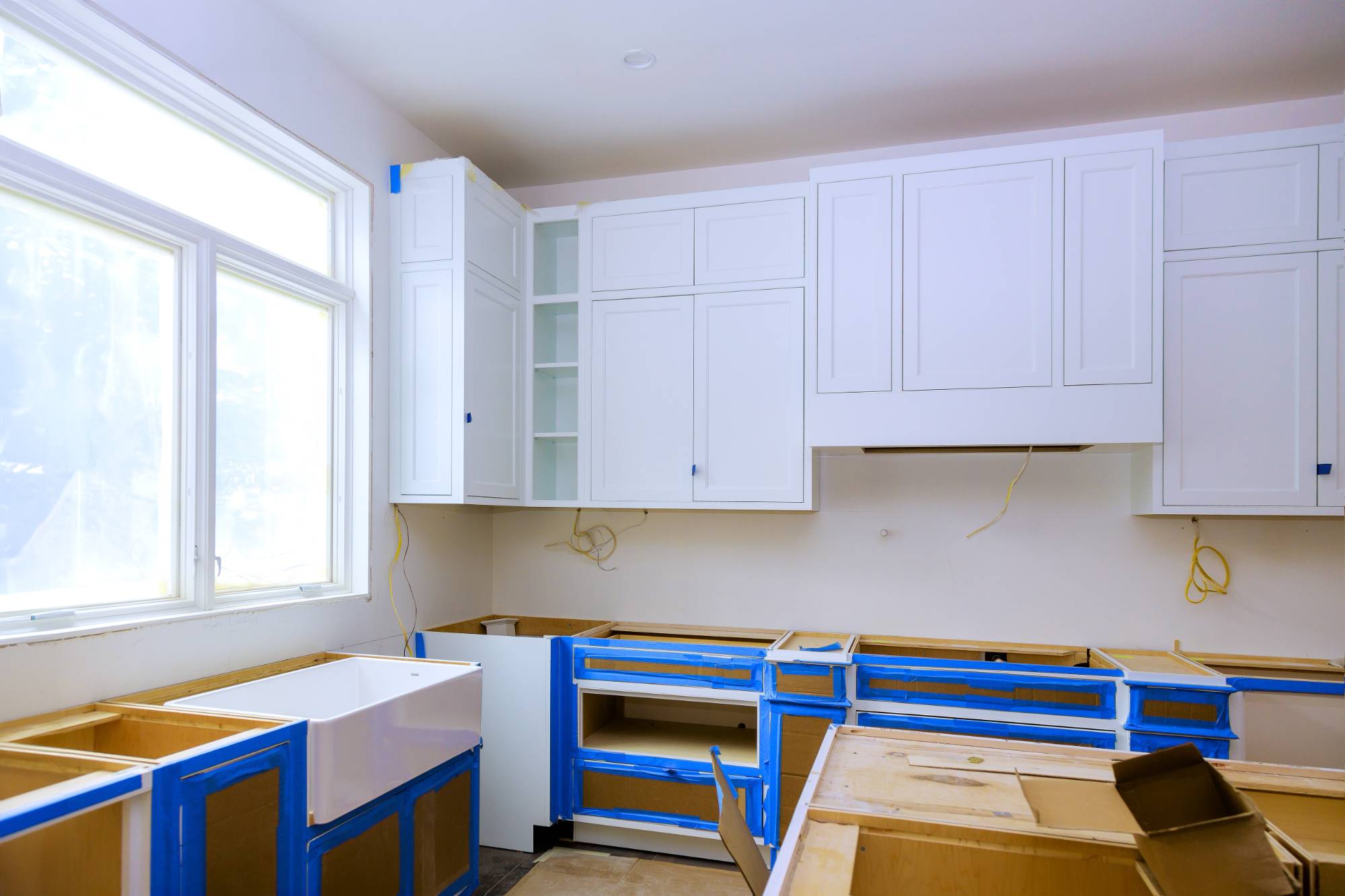 A kitchen under renovation in MI with white upper cabinets, blue protective tape on lower cabinet frames, a white farmhouse sink, exposed wires, and construction materials—perfect for a Painter Macomb & Oakland Counties, MI project.
