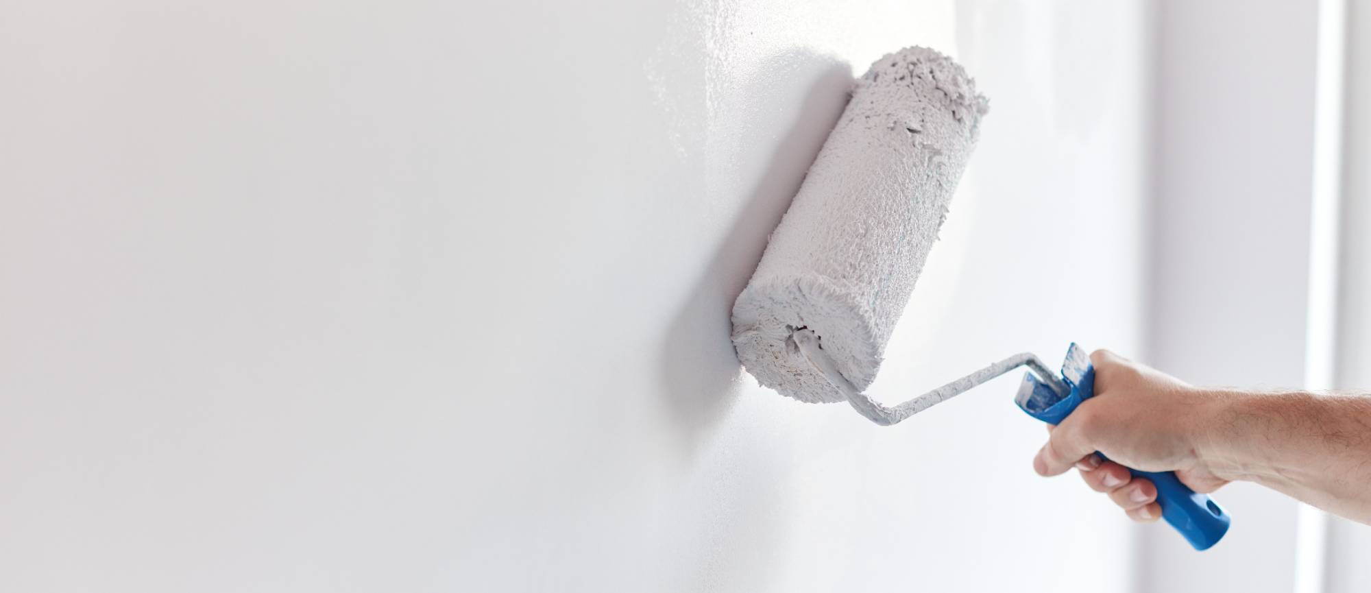A hand holds a paint roller applying white paint to a smooth wall, creating an even, fresh coat. The background is bright and minimalistic—perfect work from a skilled painter in Macomb & Oakland Counties, MI.