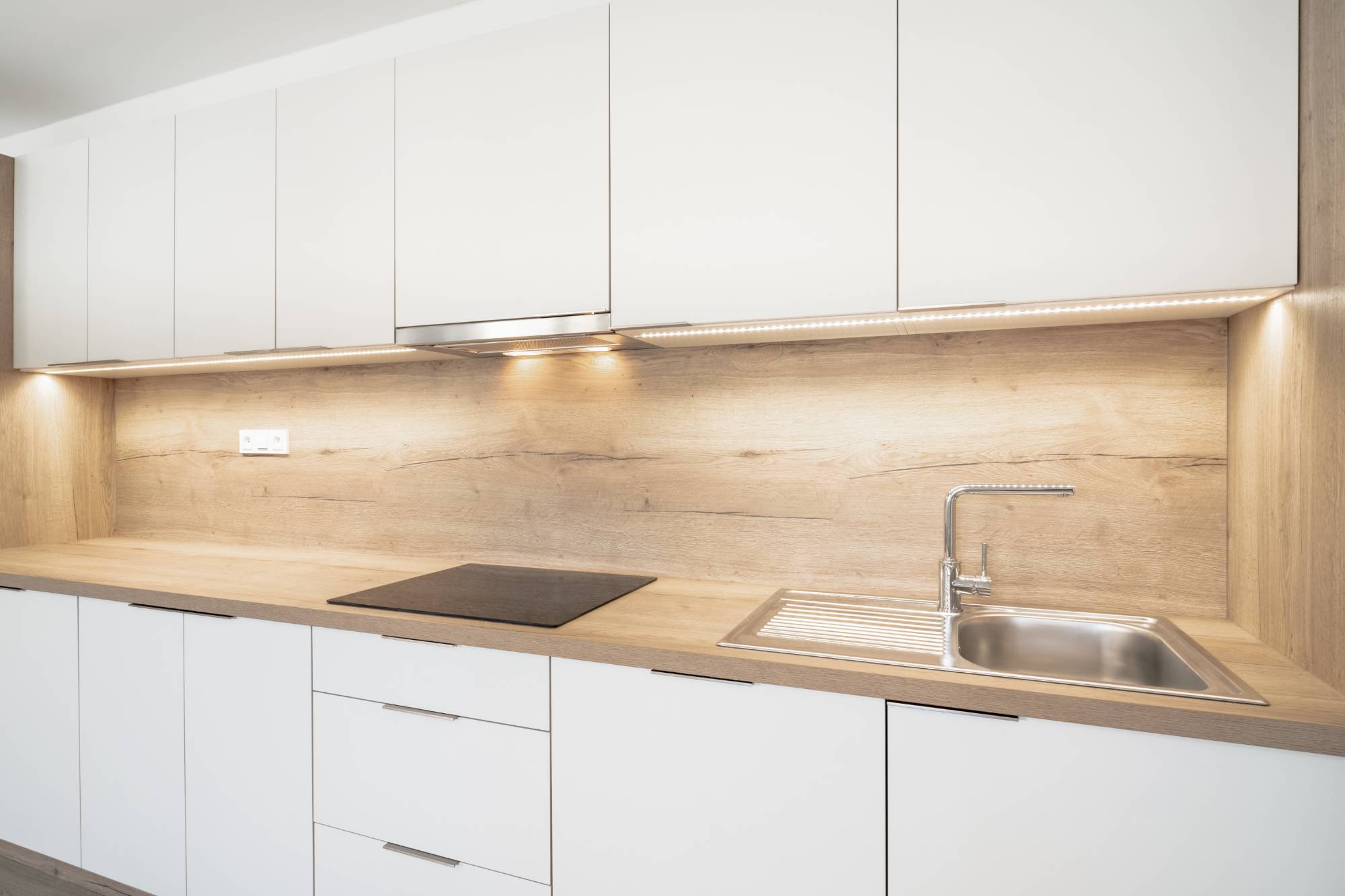 Modern kitchen with white cabinets, wooden countertop and backsplash, electric cooktop, and stainless steel sink. Under-cabinet lighting illuminates the workspace. For a fresh look, consider painting services Macomb & Oakland Counties, MI.
