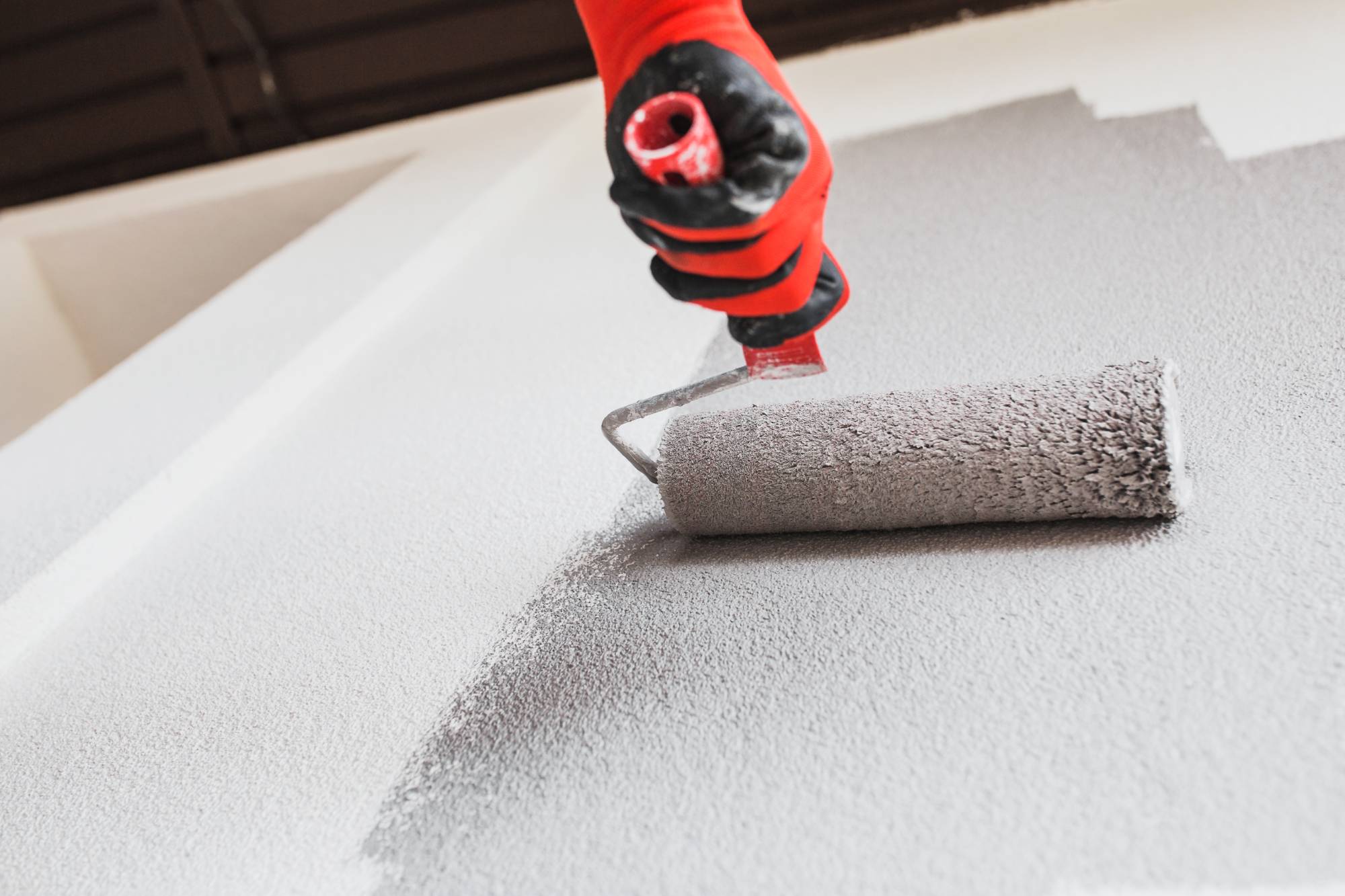A person wearing a red glove uses a paint roller to apply light gray paint to a white wall, showcasing the quality work of painting services Macomb & Oakland Counties.