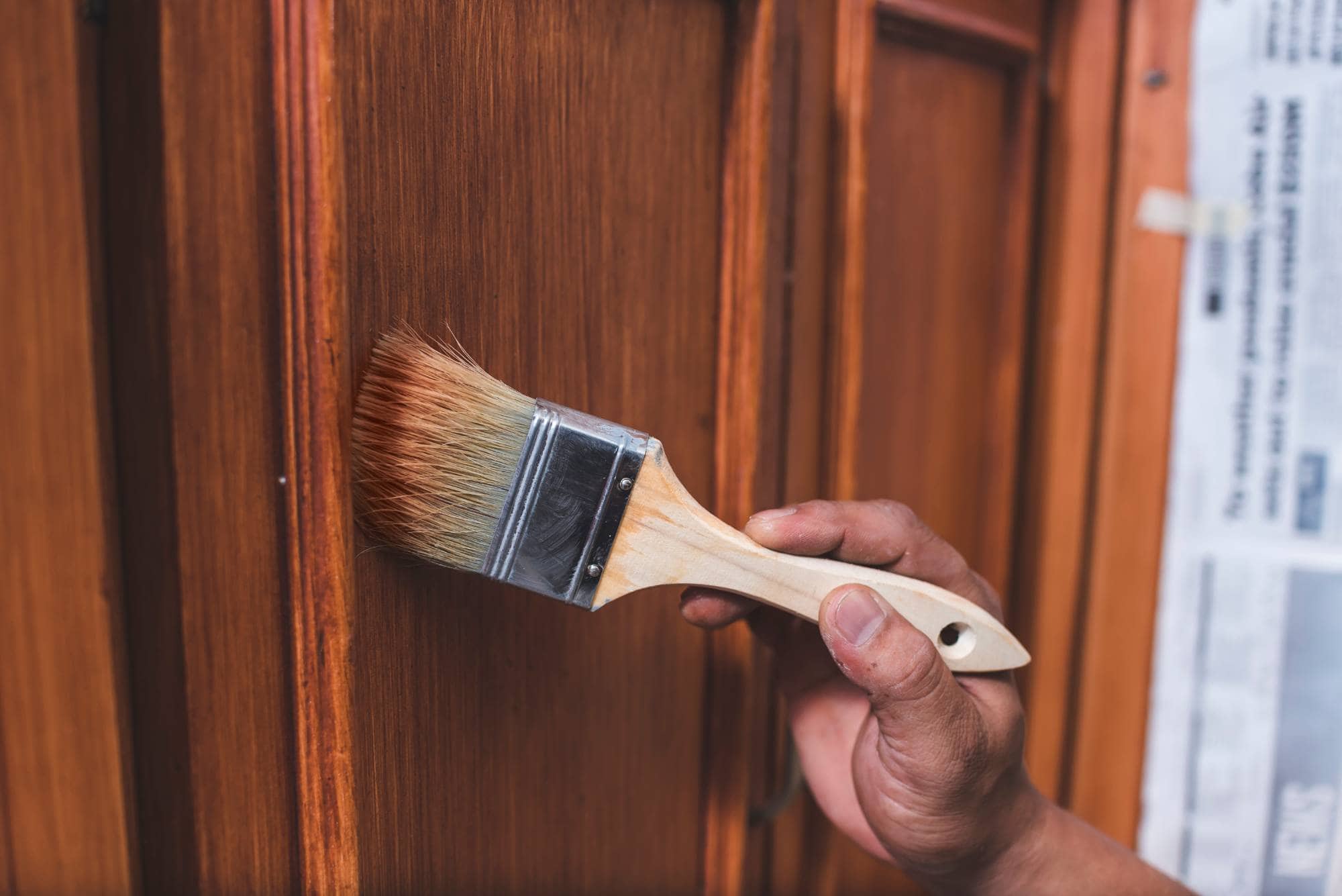 A hand holding a paintbrush applies varnish or paint to a wooden door with raised panels; part of a newspaper is visible taped to the side for protection—painting services Macomb & Oakland Counties bring care and detail to every project.