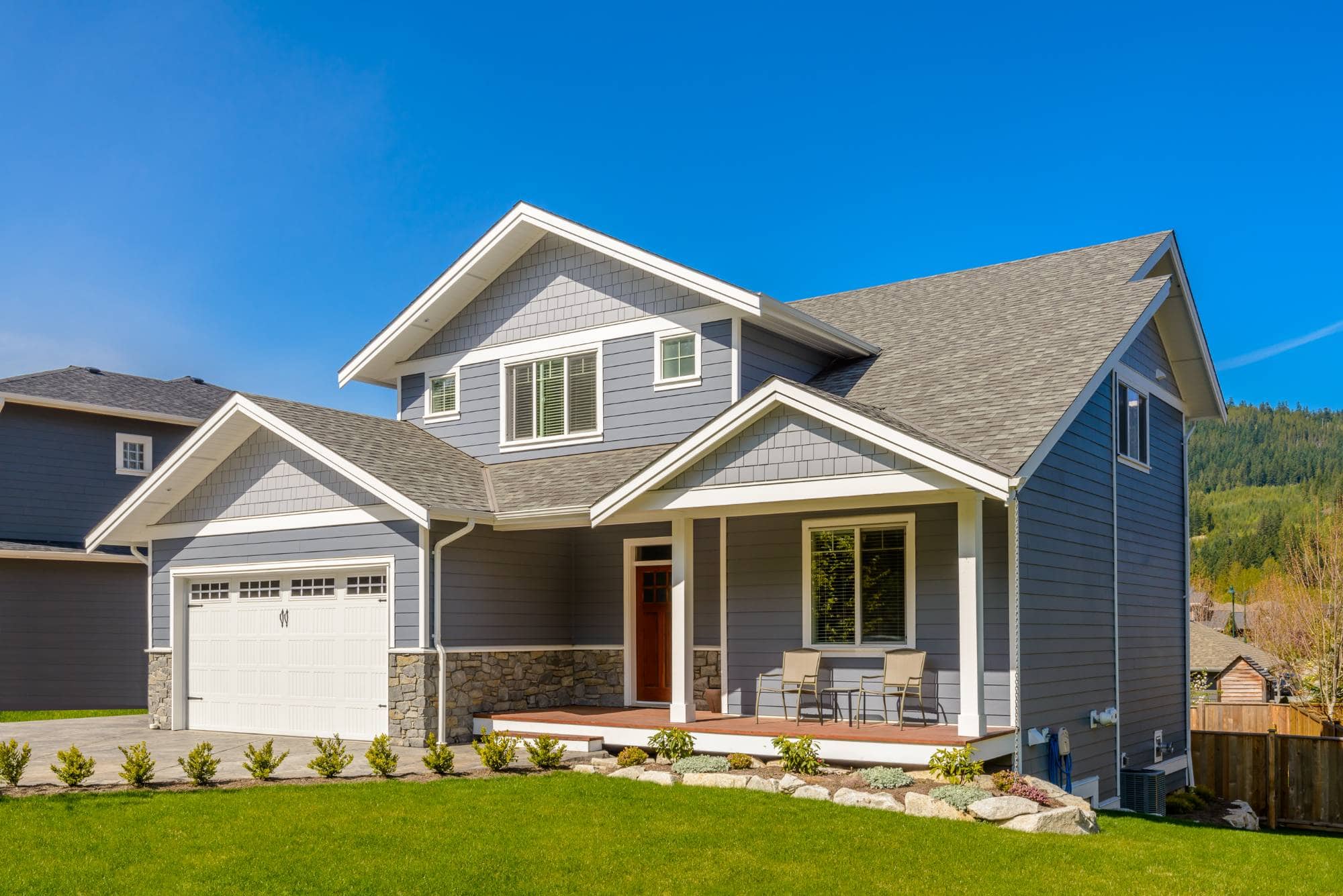 A modern two-story house with gray siding, white trim, and a two-car garage sits on a manicured lawn under a clear blue sky—expertly finished by Painter Macomb & Oakland Counties, MI.
