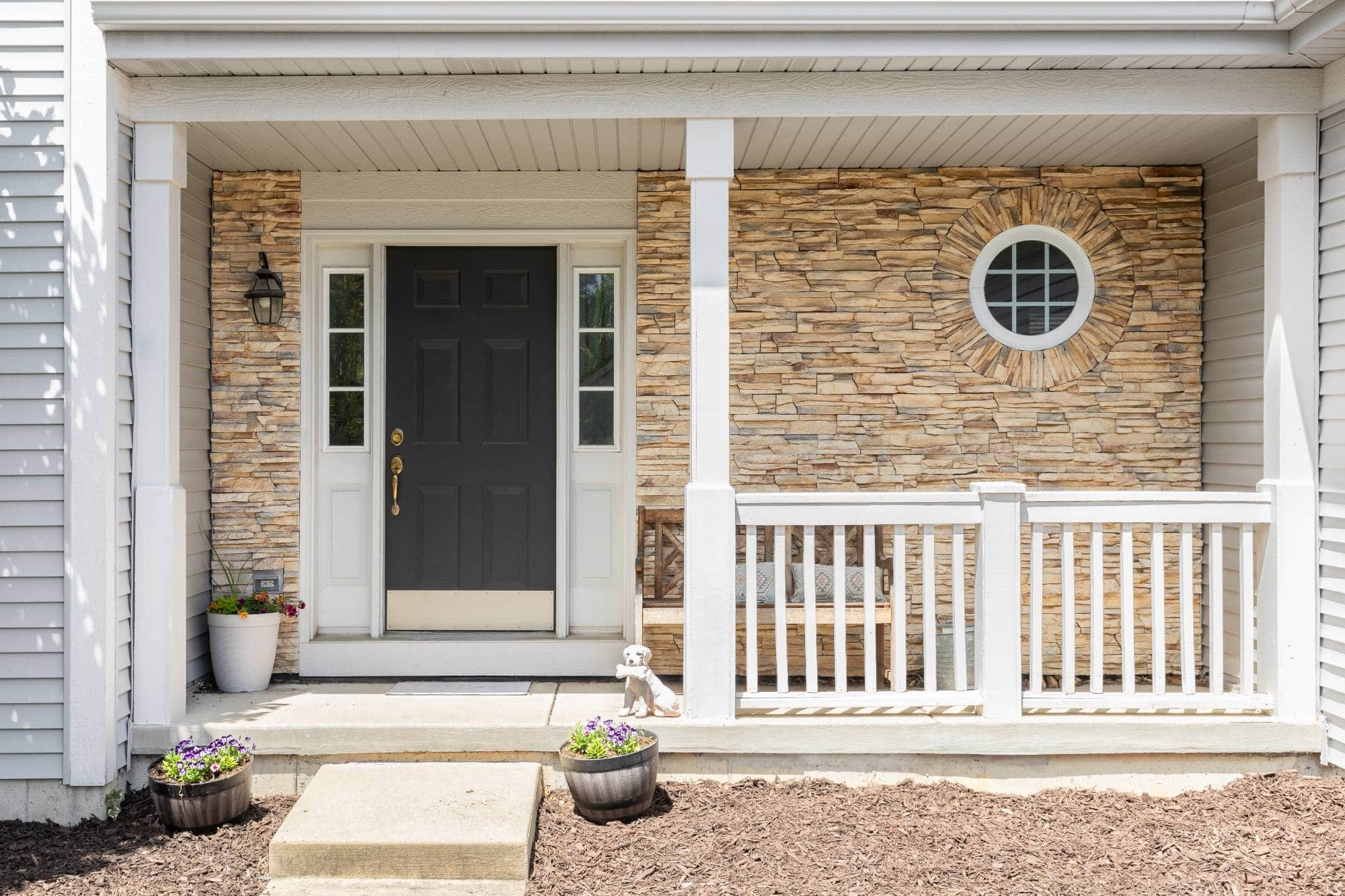 A front porch in MI with a black door, stone wall, white railing, and round window. Two potted flowers and a small dog statue sit on the steps, with a bench and more flowers—perfect for painting services Macomb & Oakland Counties, MI.
