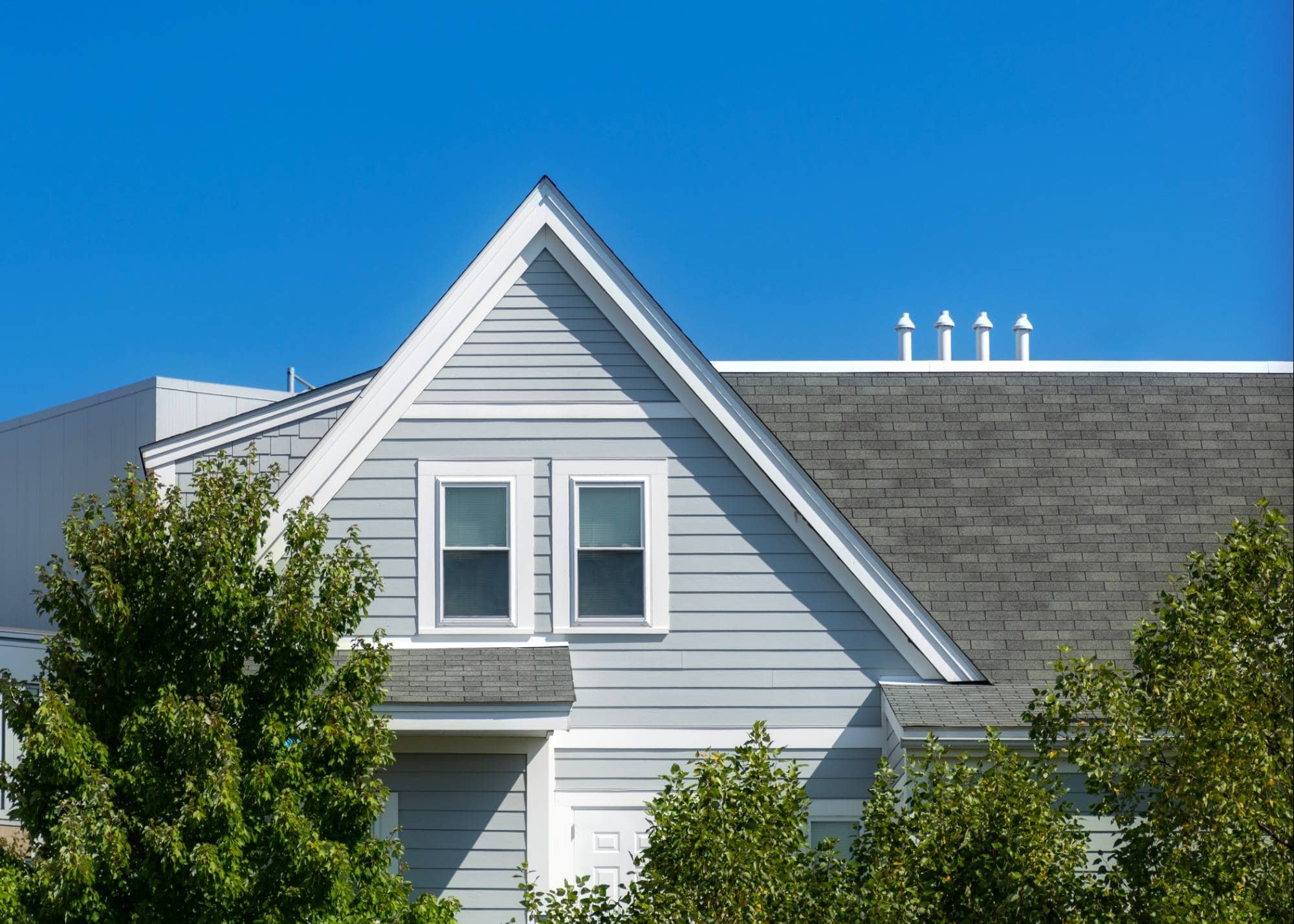 A light gray house with white trim and a steep gabled roof is partly obscured by leafy green trees. The sky above is clear and bright blue. Trust painting services Macomb & Oakland Counties, MI for expert care on homes like this.