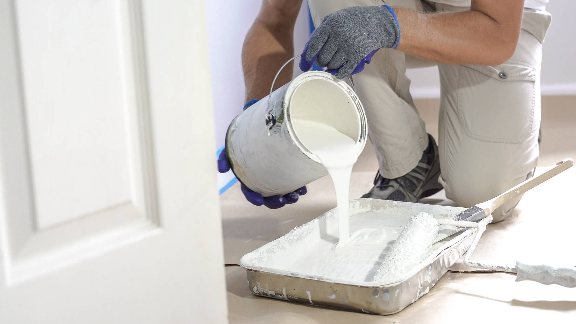 A person wearing gloves and work clothes pours white paint from a can into a paint tray, preparing to use a roller for painting. A door and part of a paintbrush are visible nearby.