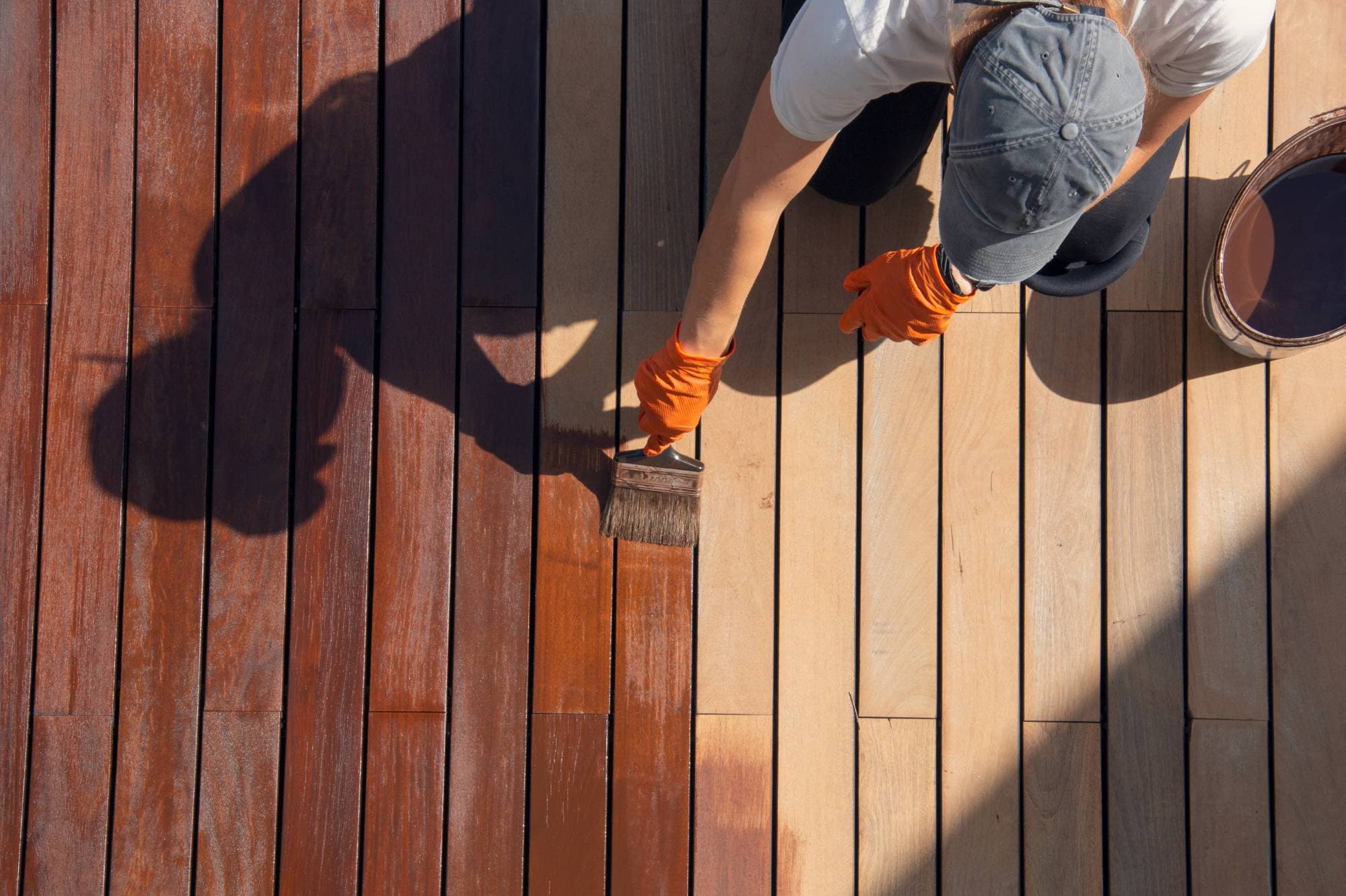 A person wearing an orange glove and gray cap applies wood stain with a brush to a wooden deck, changing the boards from light to a rich, dark brown color. A can of stain sits nearby.
