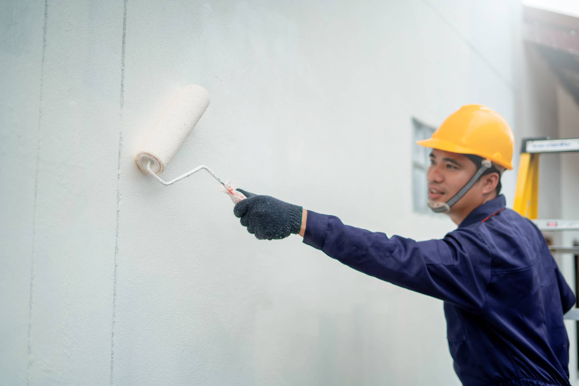 A person wearing a yellow hard hat, dark coveralls, and gloves uses a paint roller to paint a white wall. A ladder is visible in the background.