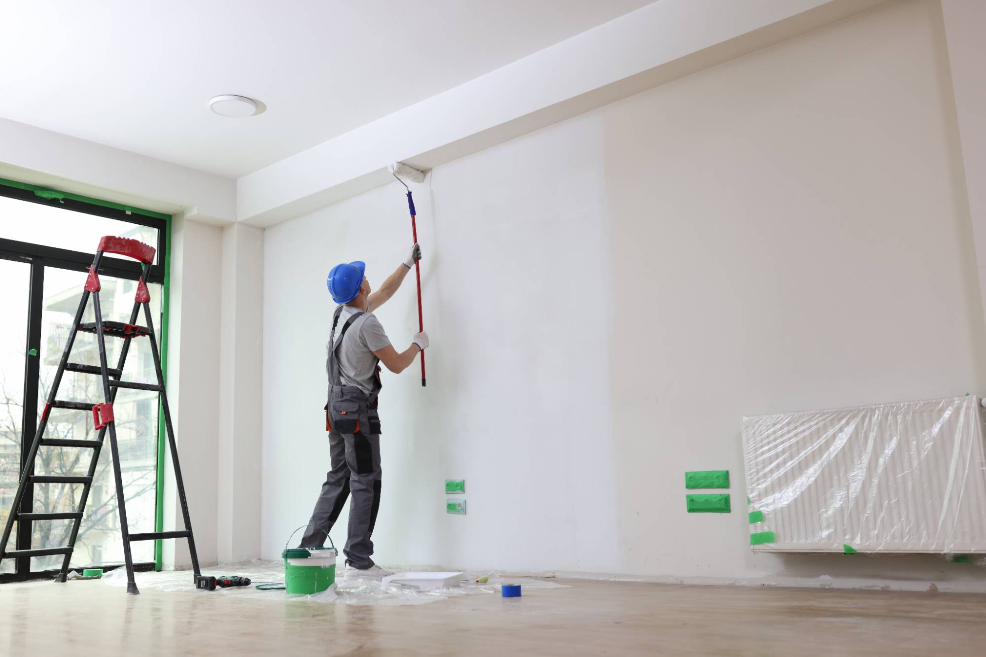 A person wearing a blue hard hat and work clothes uses a roller to paint a high wall white. A red step ladder, green tape, a bucket, and a covered radiator are visible in the bright, modern room.