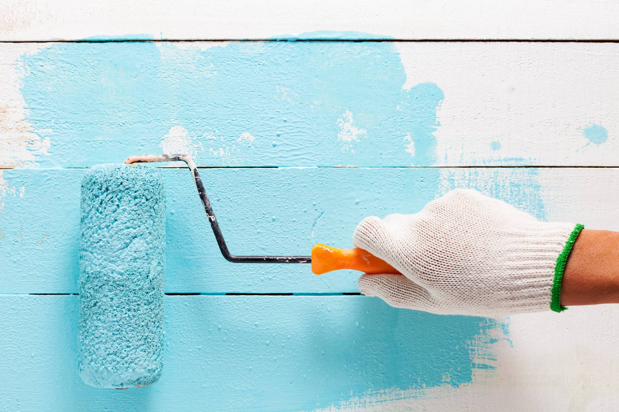 A hand in a white glove holds a paint roller with blue paint, rolling it over white wooden boards, partially covering them with a fresh coat of blue paint.