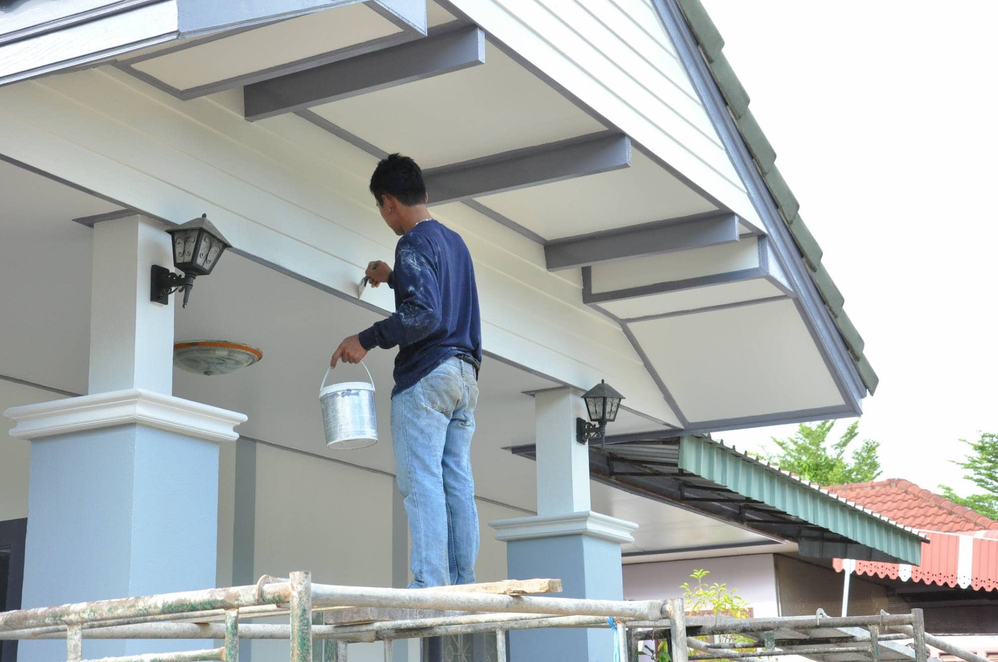 A man standing on scaffolding paints the exterior ceiling of a house white, holding a paint bucket in one hand and a brush in the other. He works near two wall-mounted outdoor lights.