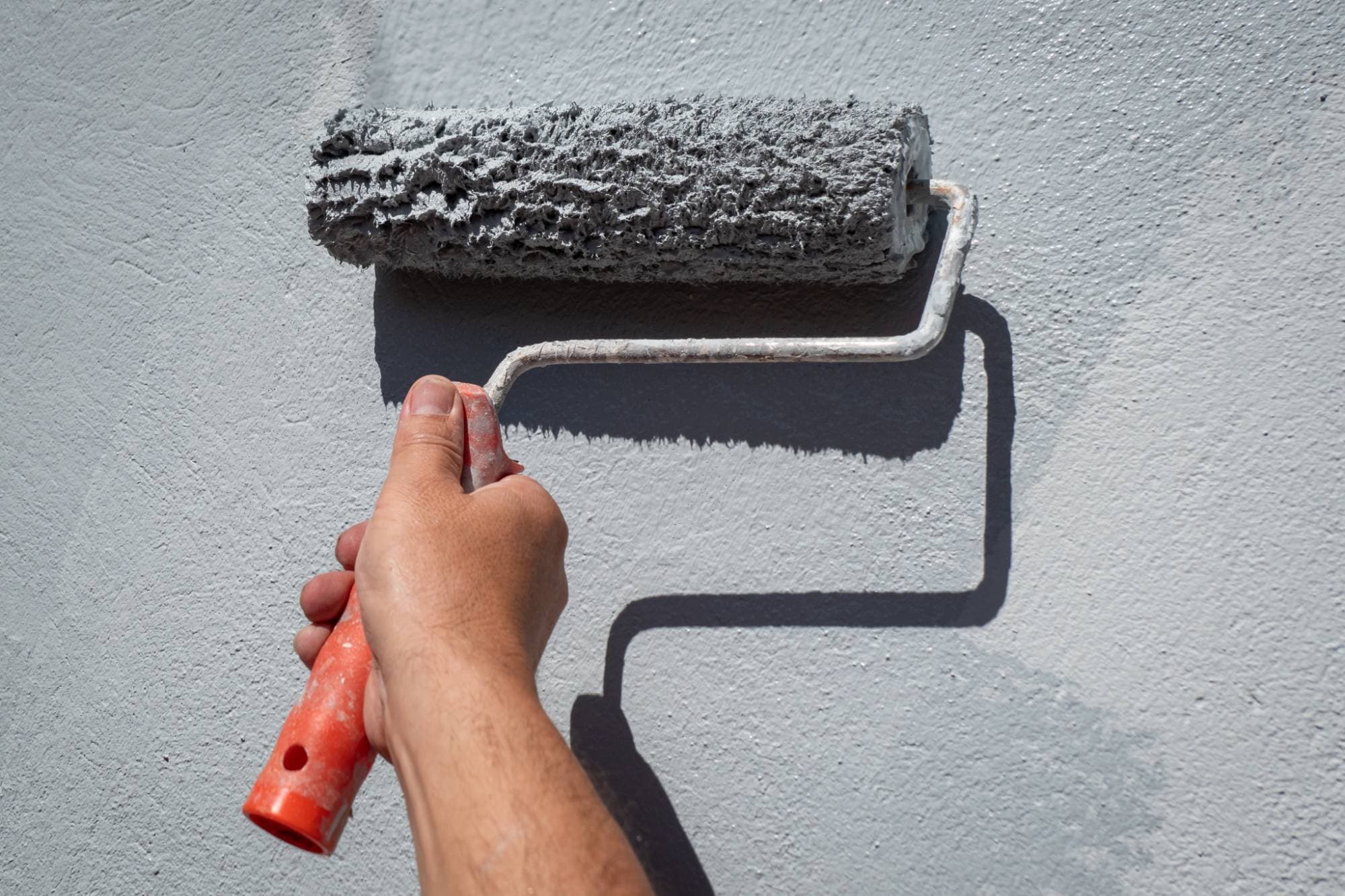 A hand holds an orange-handled paint roller, applying light gray paint to a textured wall. The roller is covered in fresh paint, and the wall appears partially painted.