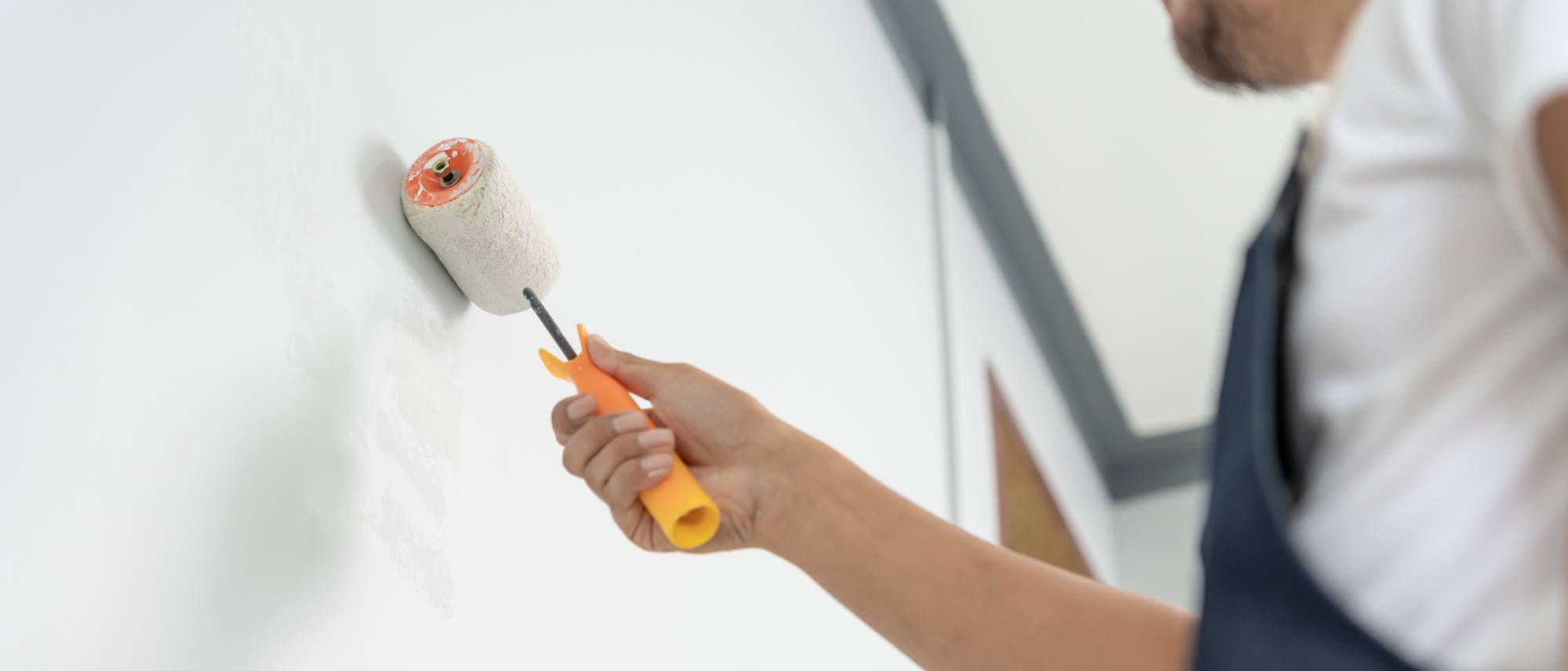 A person in a white shirt and dark apron uses a paint roller to apply white paint to a wall. The focus is on their arm and the roller, with the background slightly blurred.