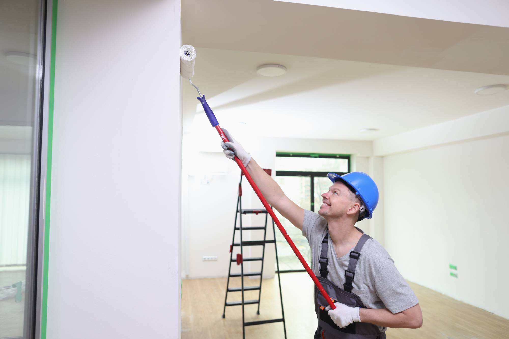 A worker wearing a blue hard hat and overalls uses a paint roller with an extension pole to paint a white interior wall. A ladder and large windows are visible in the background of the bright room.