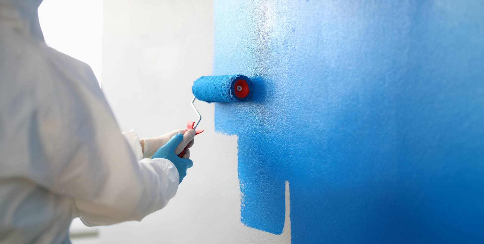 A person wearing protective gloves and white clothing is painting a wall bright blue using a paint roller. The fresh blue paint contrasts with the unpainted white area of the wall.