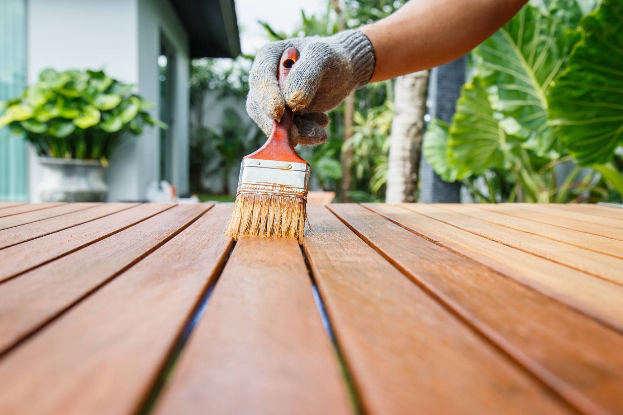 A gloved hand holds a paintbrush, applying varnish to a wooden outdoor surface. Green plants and a house are visible in the blurred background.