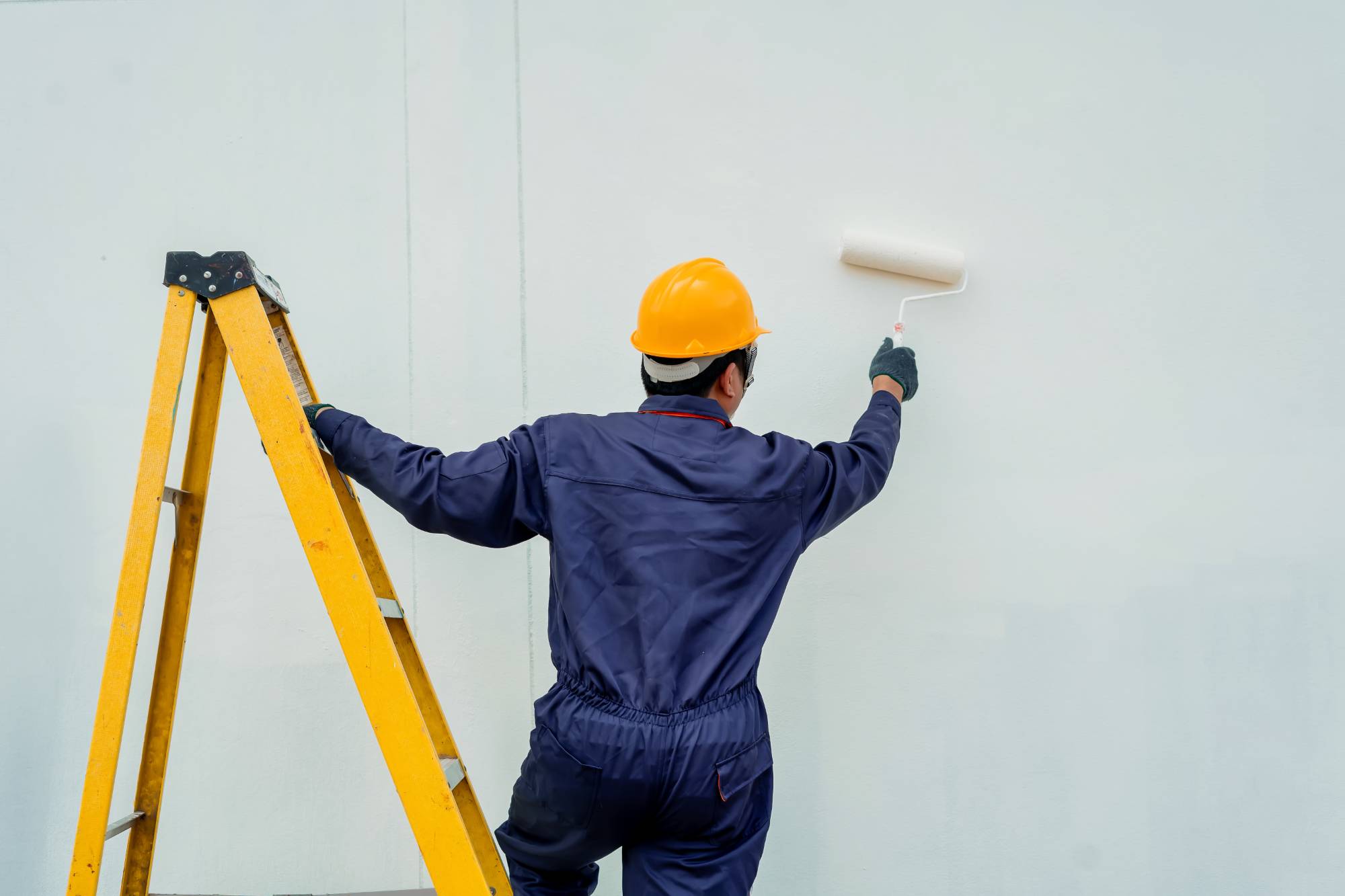 A worker wearing a yellow hard hat and blue coveralls uses a paint roller to paint a white wall, standing next to a yellow ladder.