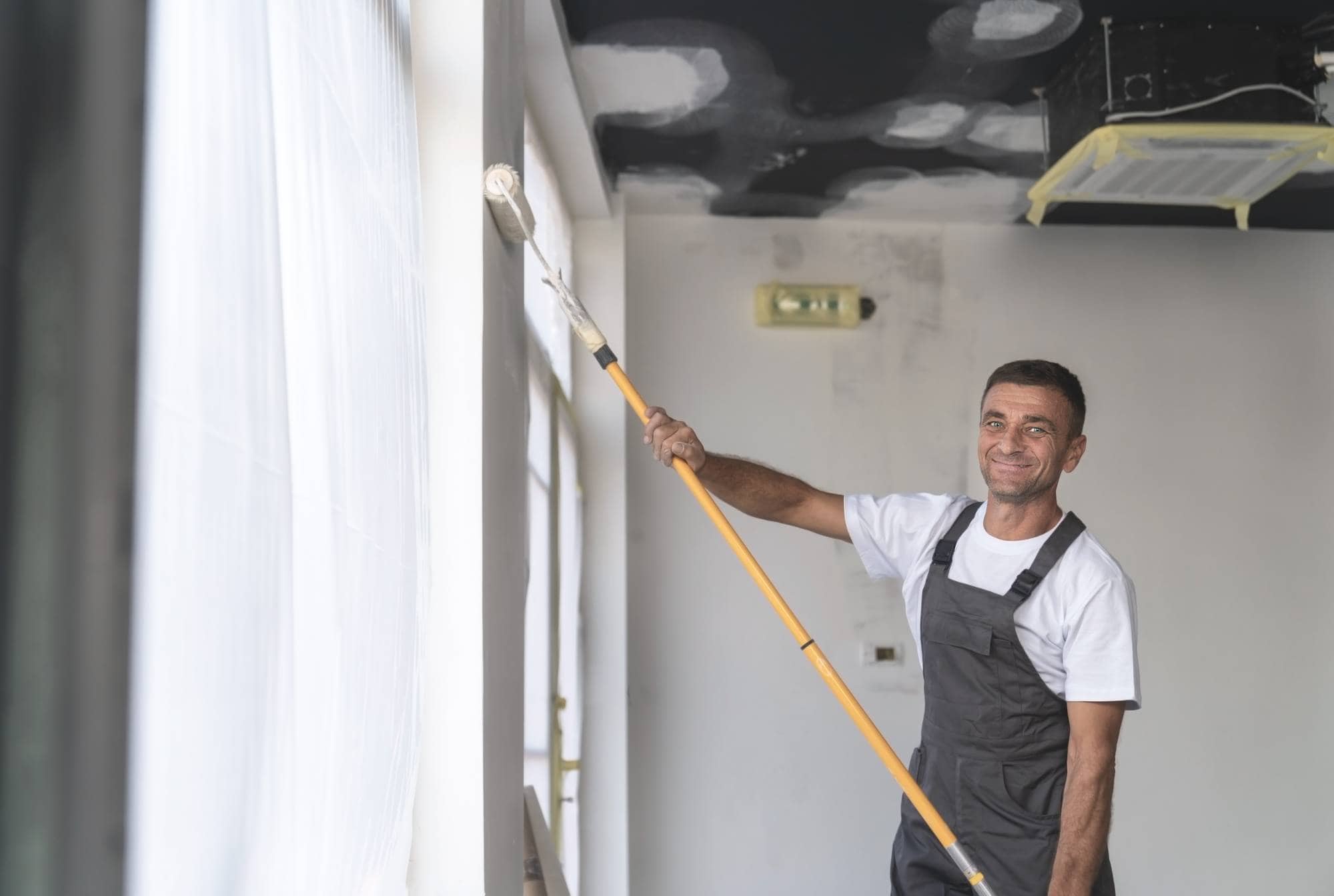 A man wearing overalls and a white t-shirt uses a long-handled roller to paint a white wall inside a room. He is smiling, and the ceiling above him appears unfinished.