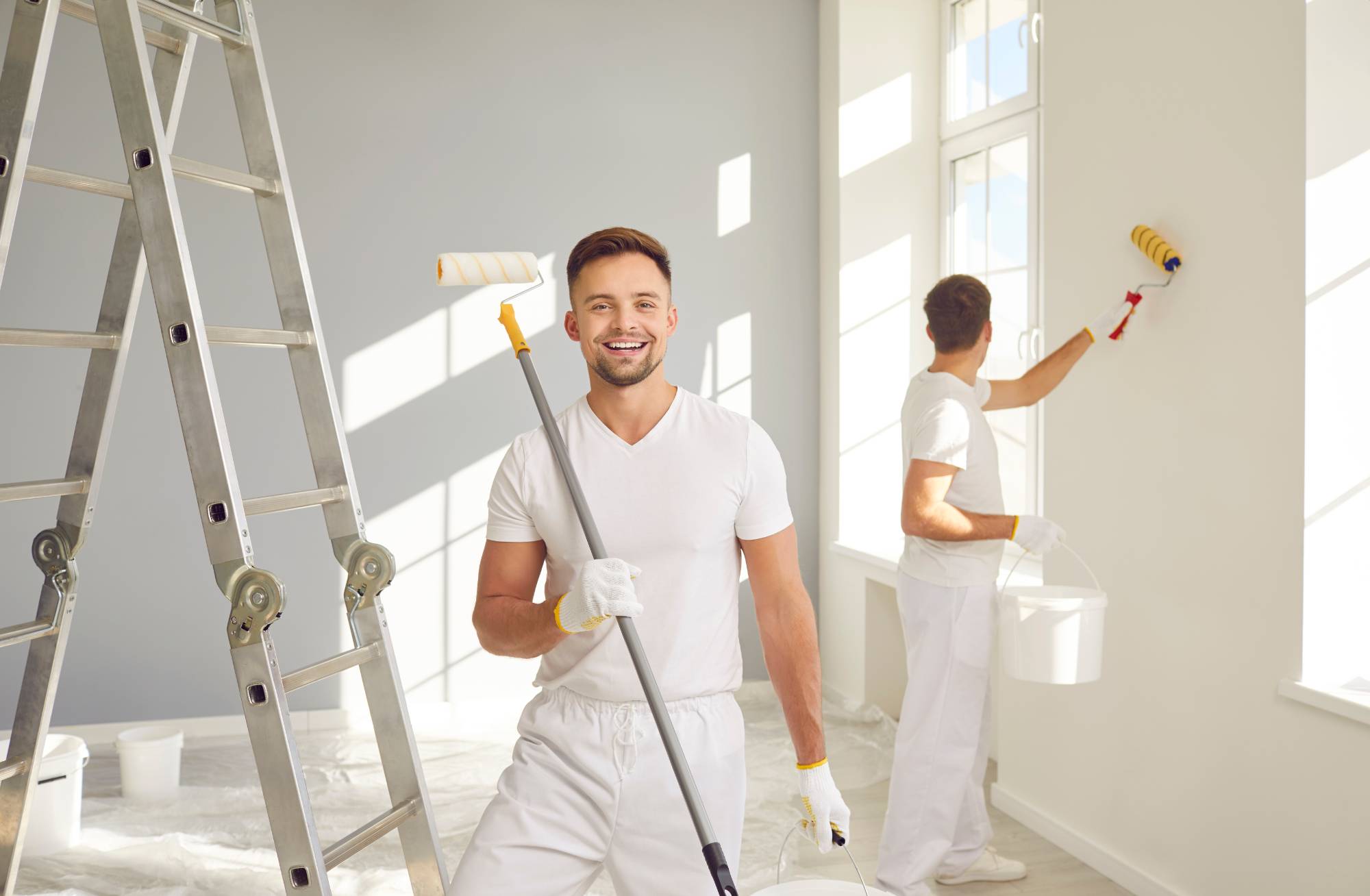 Two men in white clothes painting a bright room; one smiles at the camera holding a paint roller, while the other paints a wall near a window. A ladder and painting supplies are visible.