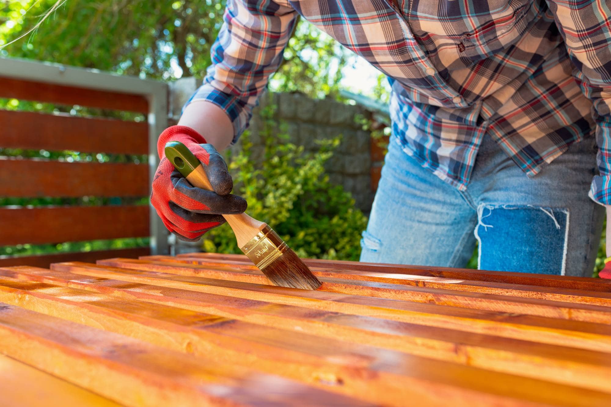 A person wearing gloves and a plaid shirt paints a wooden outdoor surface with a brush, applying a protective finish. Green foliage and a wooden bench are visible in the background.