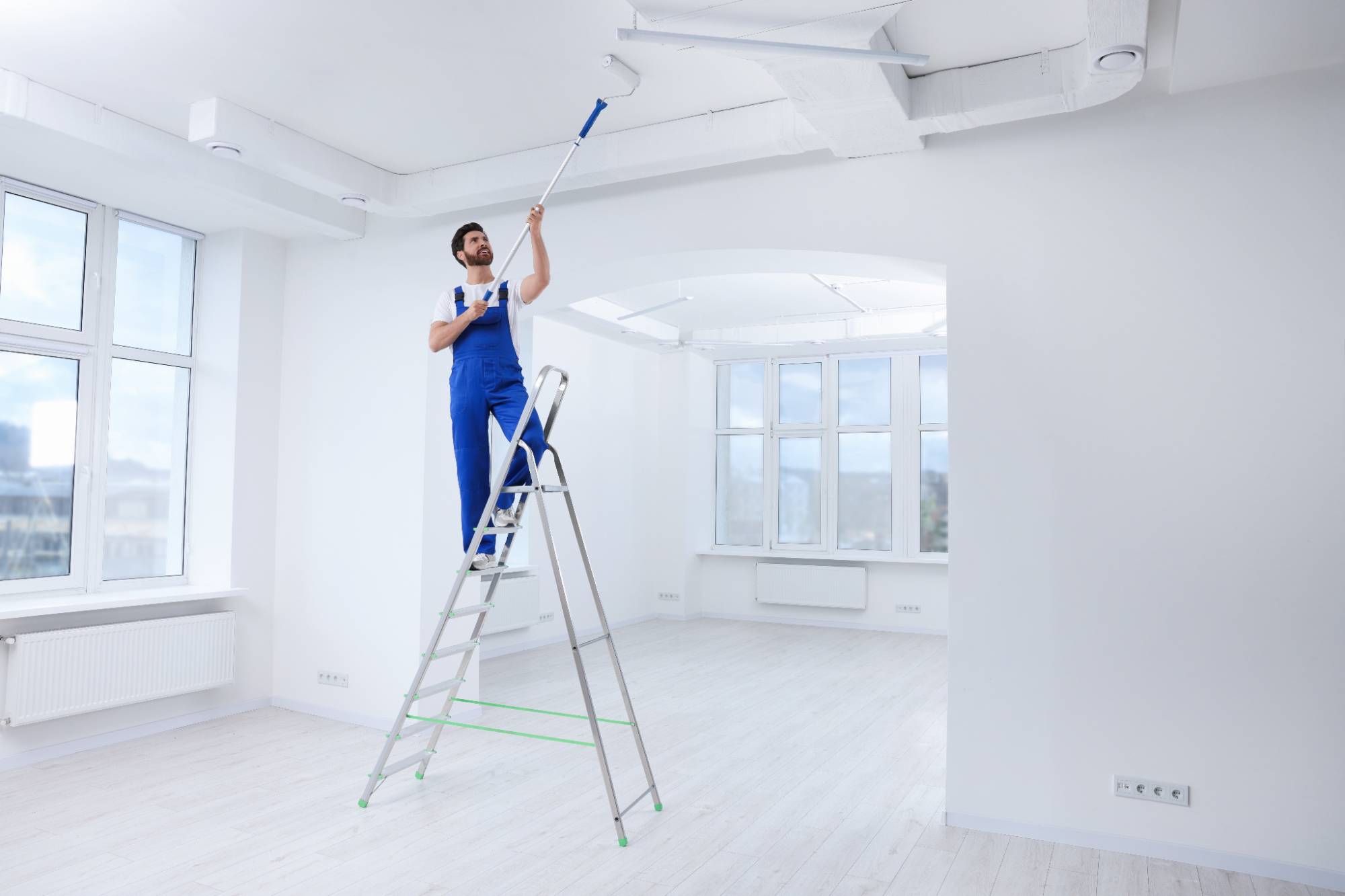 A person in blue overalls stands on a ladder, painting a white ceiling with a roller in a bright, empty room with large windows and light-colored floors.