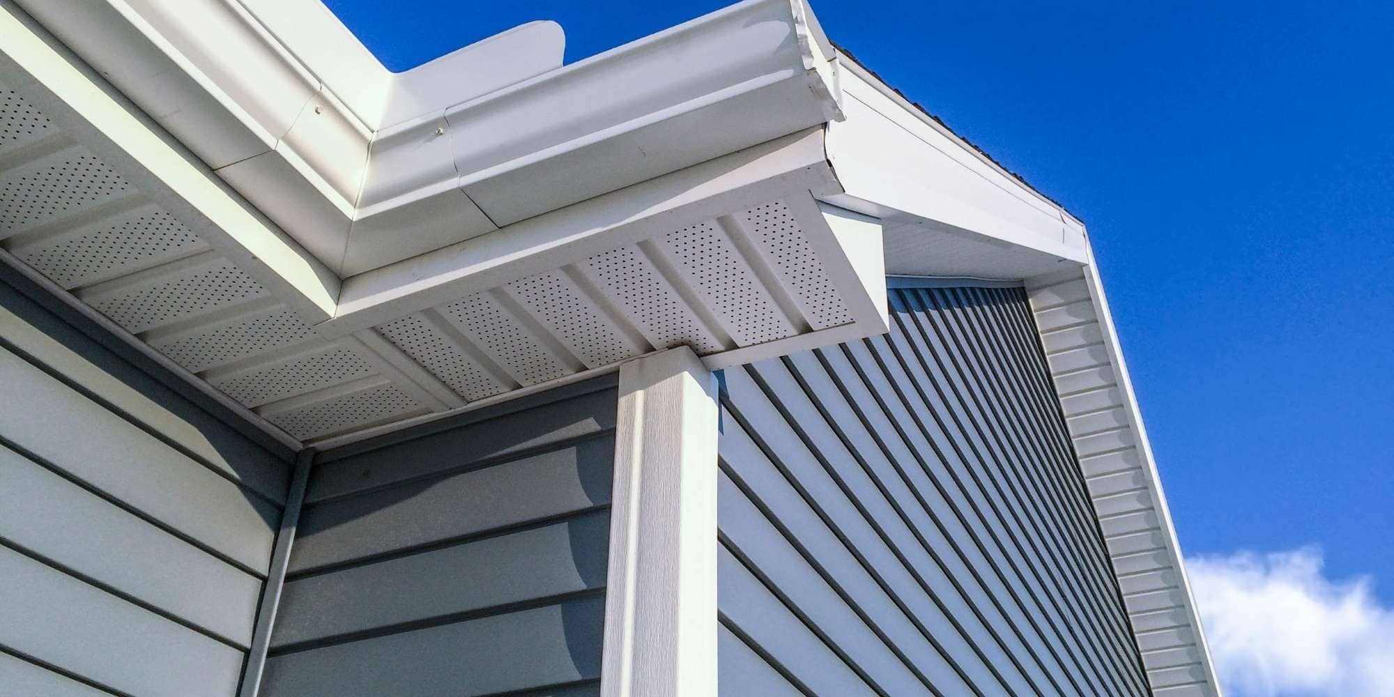 Close-up view of the corner of a house in MI with light gray vinyl siding, white soffits, and trim, set against a clear blue sky—expertly finished by Painter Macomb & Oakland Counties, MI.
