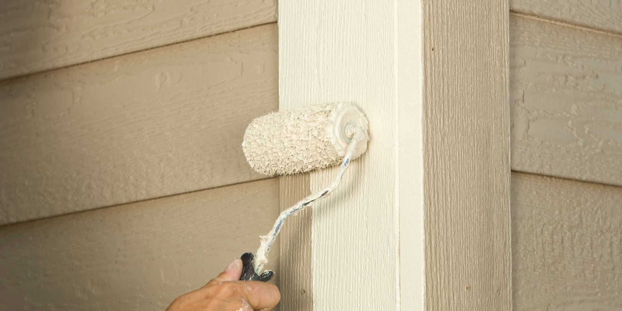 A hand uses a paint roller to apply light-colored paint to the exterior siding and corner of a house, showcasing the textured wood grain pattern. Trust Painter Macomb & Oakland Counties for expert painting services in MI.