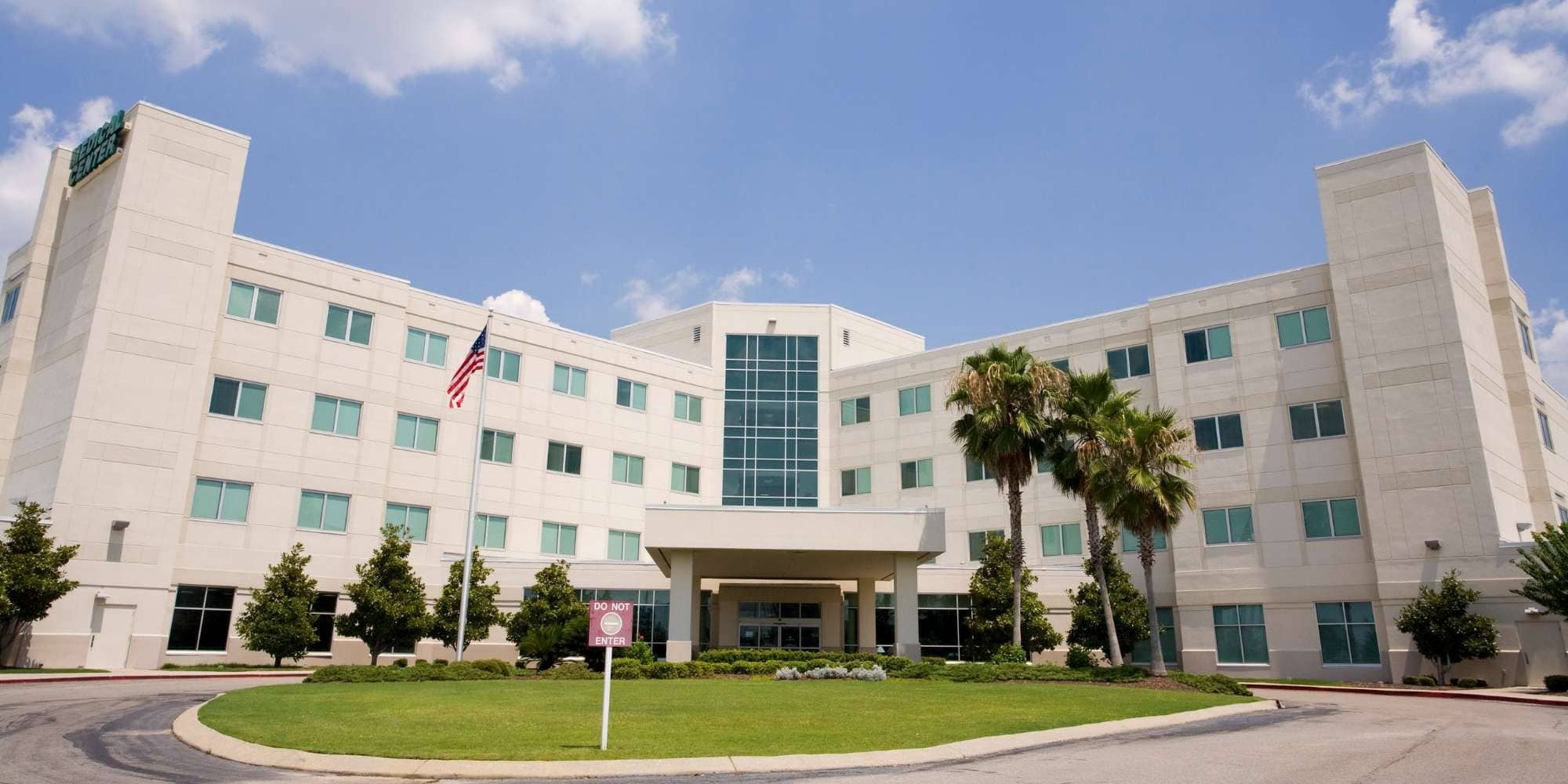 A large, modern hospital building with four stories, an American flag outside, palm trees, and expertly maintained landscaping under a blue sky—showcasing quality painting services Macomb & Oakland Counties trusts.