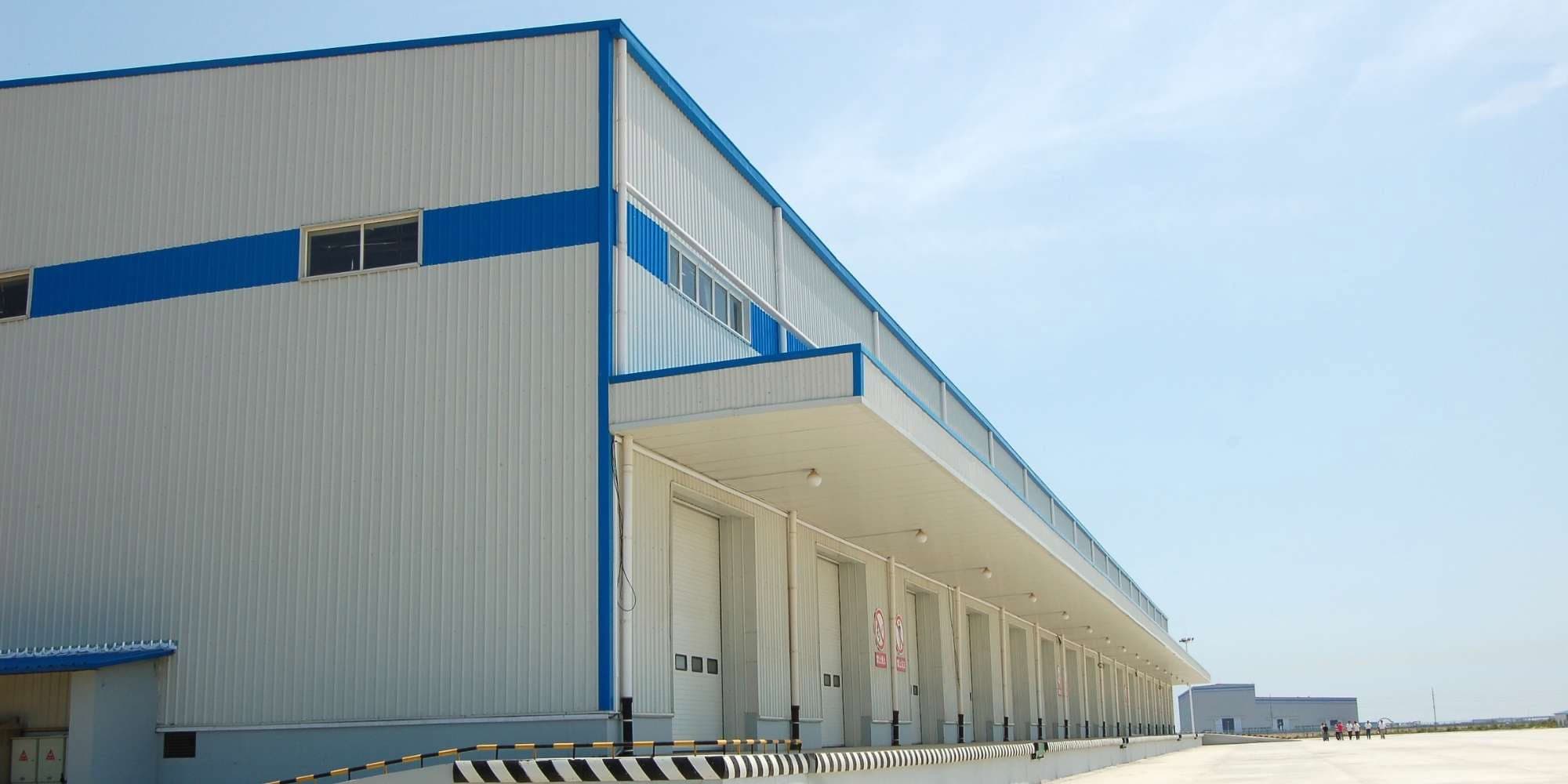 A large, modern warehouse with multiple loading docks and a blue and white exterior, freshly finished by painting services Macomb & Oakland Counties. The area in front is paved and spacious, ideal for trucks under a clear sky.