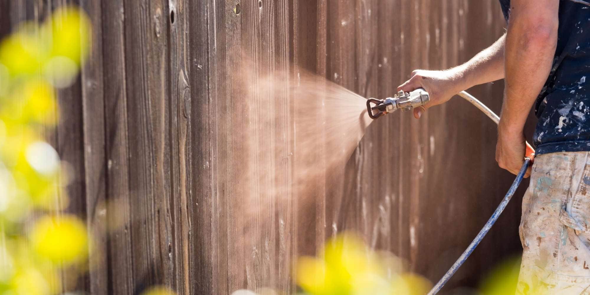 A person sprays paint onto a wooden fence using a paint sprayer, showcasing the expertise of painting services Macomb & Oakland Counties, MI. The person’s arm is visible in a dark shirt with paint splatters, as greenery frames the scene.