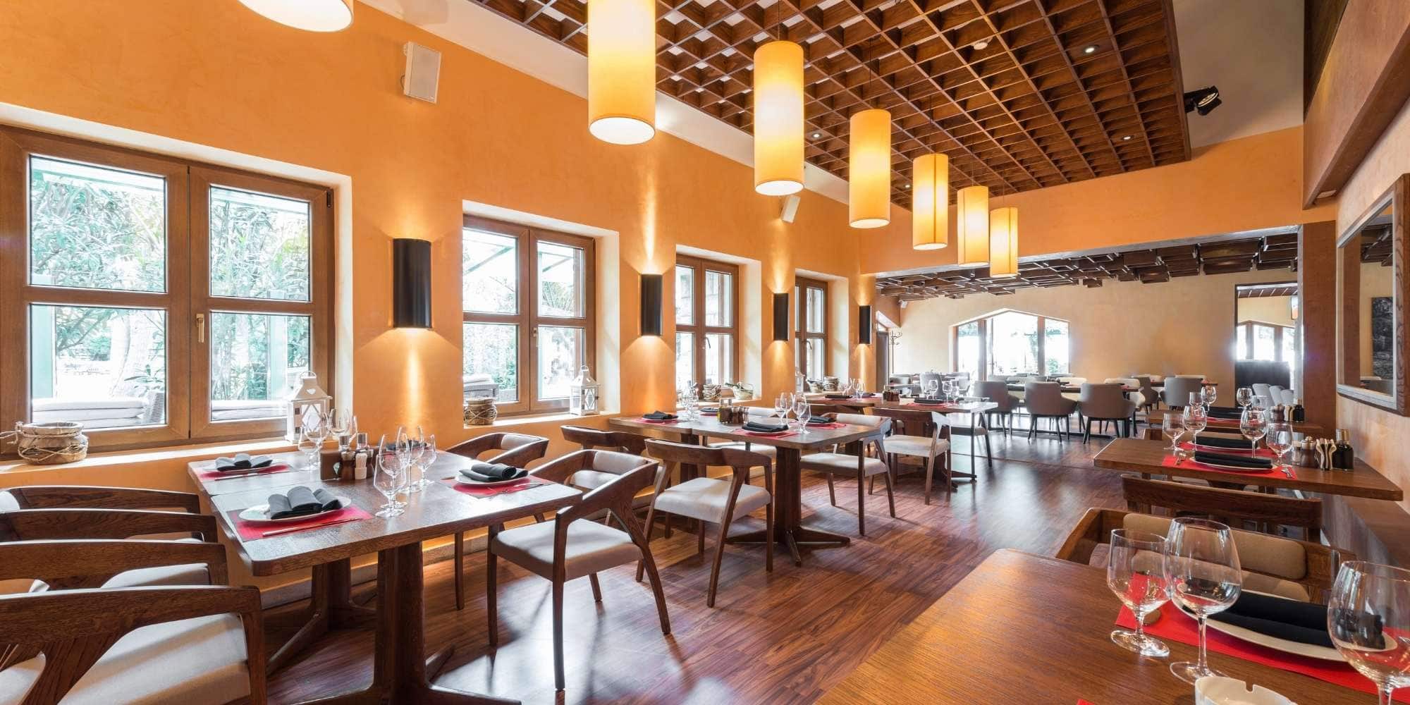 A modern restaurant interior with wooden tables and chairs, set with glassware and napkins. Warm yellow walls, expertly finished by a Painter Macomb & Oakland Counties, MI, and hanging lights create a cozy, inviting atmosphere.