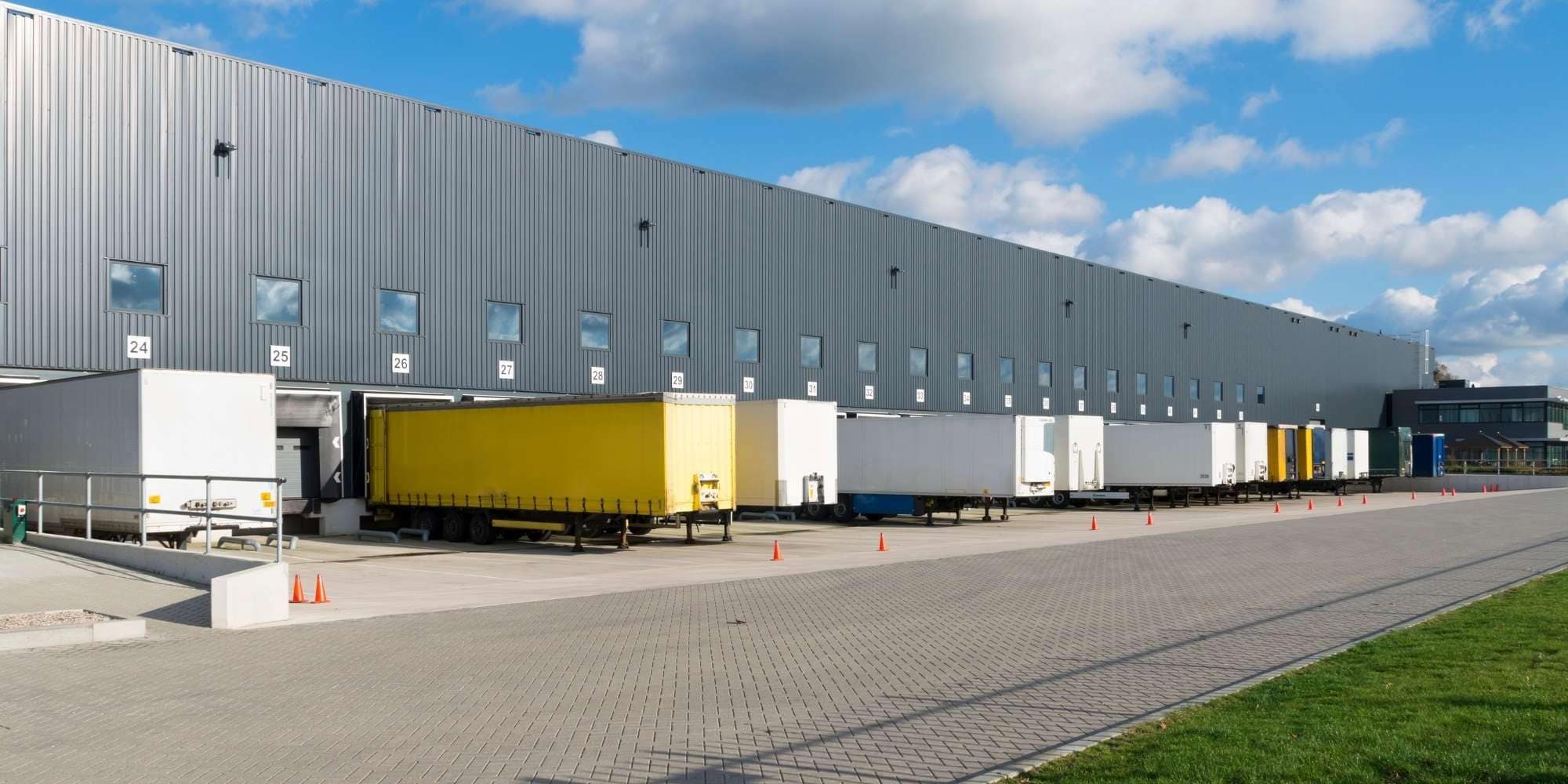 A row of loading docks at a warehouse in MI features several trailers, including a bright yellow one, parked for loading or unloading under a partly cloudy sky. The area is clean and paved, marked with some orange traffic cones.