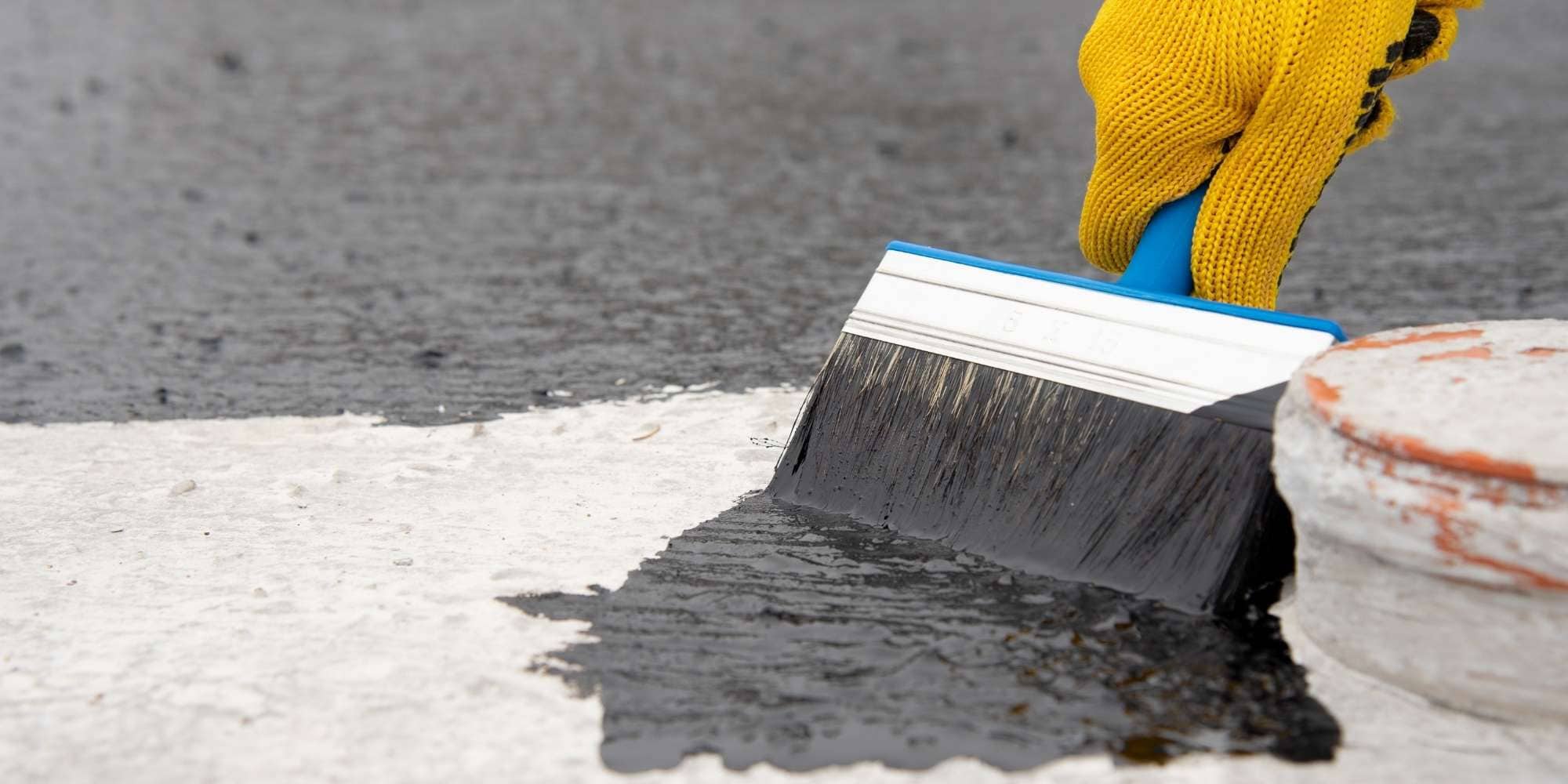 A person wearing a yellow glove applies black waterproofing sealant to a concrete surface using a wide brush, showcasing the precision you can expect from painting services Macomb & Oakland Counties, MI. Part of the concrete is already expertly coated.