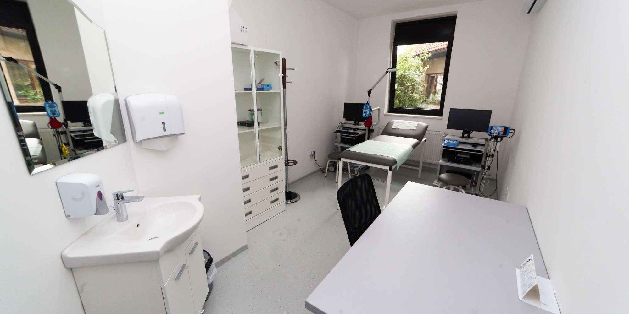 A clean, modern medical examination room in MI features a desk, chair, examination table, medical equipment, and natural light—a space that could benefit from professional painting services in Macomb & Oakland Counties for a fresh look.