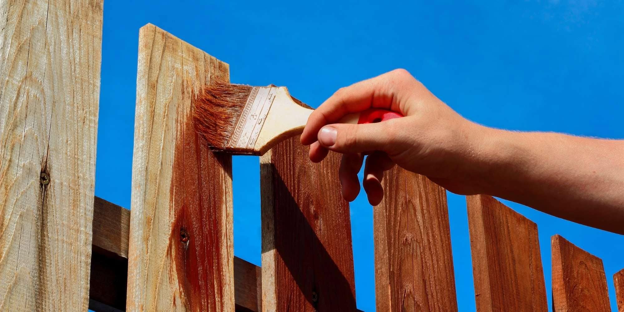 A hand uses a paintbrush to apply a brown stain to a wooden fence under a clear blue sky, showcasing expert painting services in Macomb & Oakland Counties, MI.