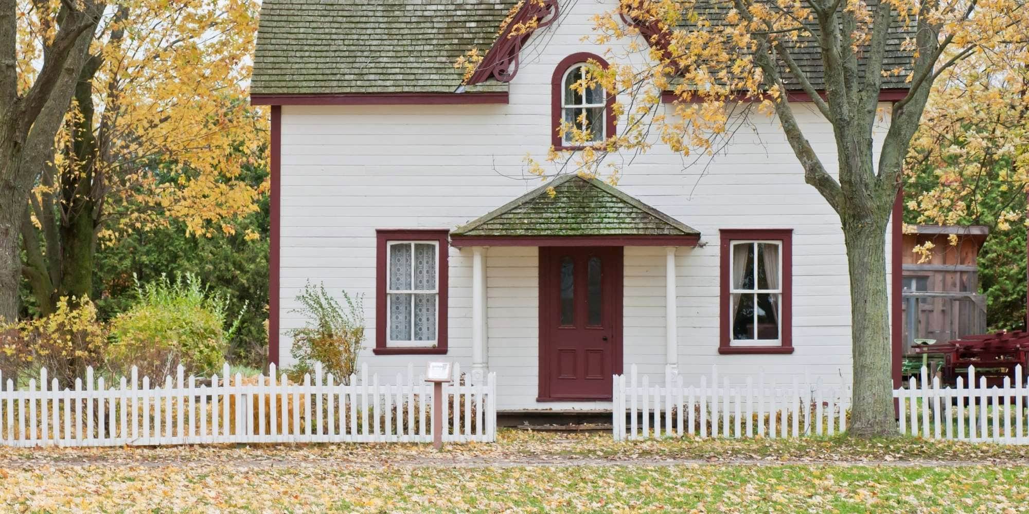 A white house with a red door and window frames sits behind a white picket fence in MI, surrounded by trees with yellow autumn leaves. Fallen leaves cover the lawn and walkway—a perfect scene for painting services Macomb & Oakland Counties.