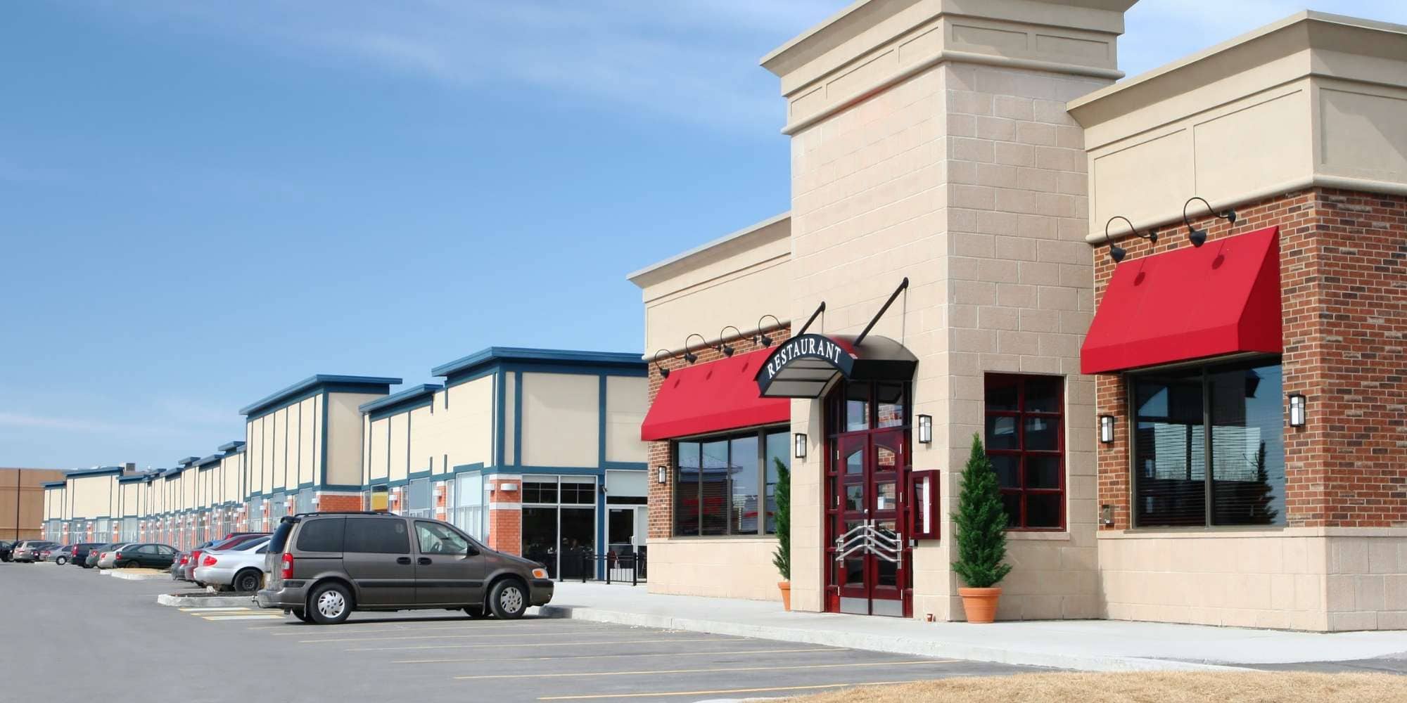 A row of modern commercial buildings with red awnings and large windows, including a restaurant, in MI—ideal for painting services in Macomb & Oakland Counties—stands beside a parking lot filled with several cars under a clear sky.