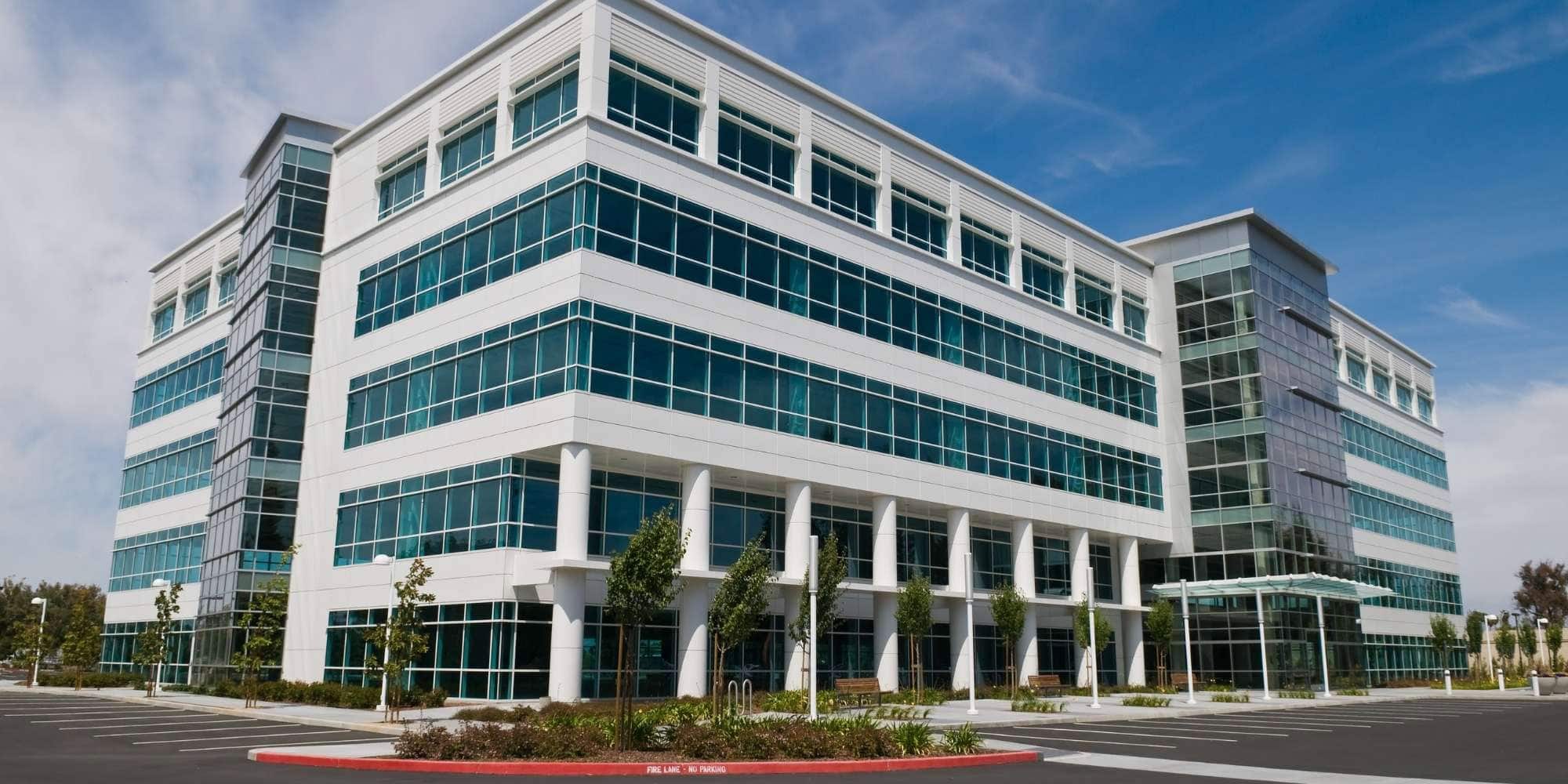 A modern, multi-story office building with large glass windows and white trim in MI, surrounded by a nearly empty parking lot and landscaped greenery under a blue sky with light clouds.