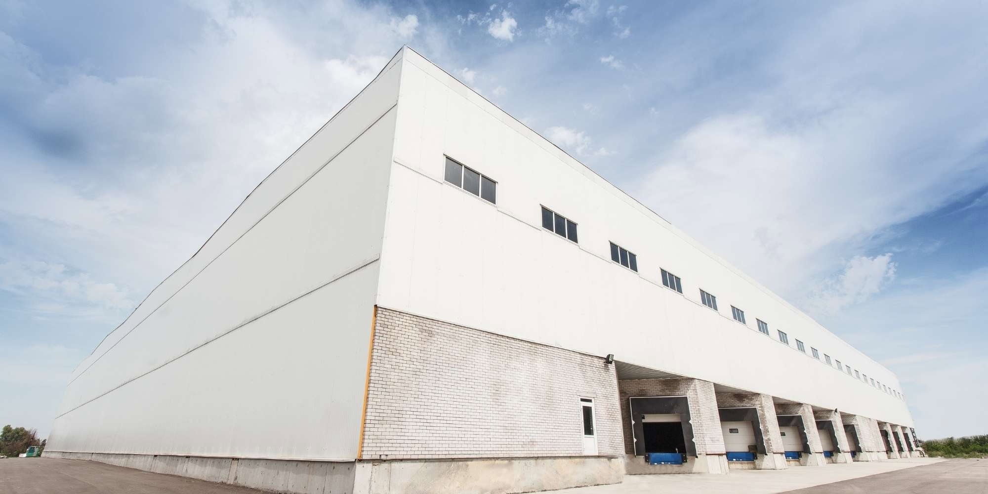 Large white industrial warehouse in MI with multiple loading bays and garage doors, seen from a low angle under a partly cloudy sky. Ideal for businesses seeking space or Painter Macomb & Oakland Counties services, with small upper windows and a spacious paved area out front.