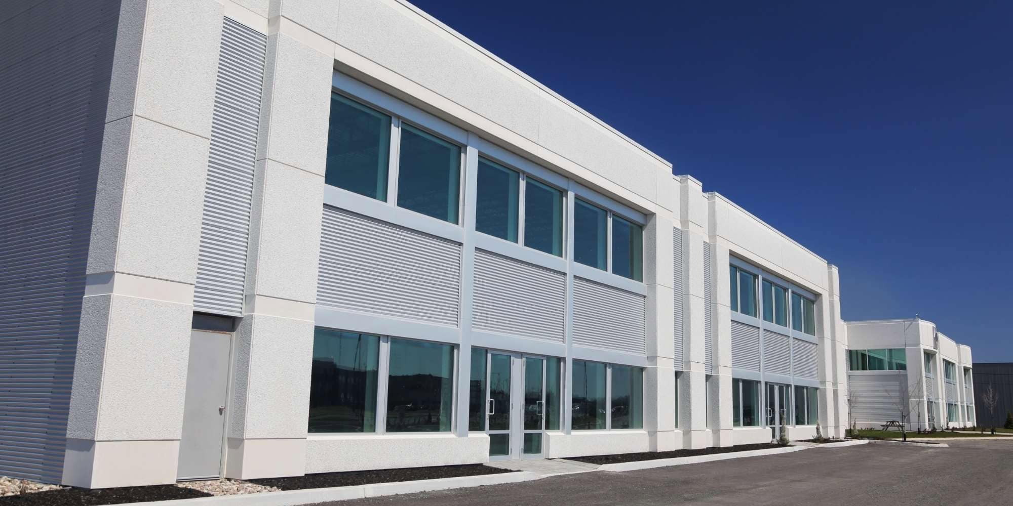 A modern, white industrial building with large glass windows and a flat roof, set against a clear blue sky in MI. There is a paved driveway and minimal landscaping—ideal for painting services Macomb & Oakland Counties.
