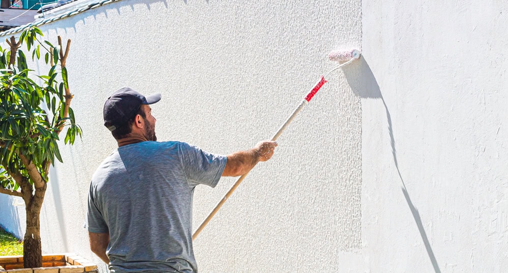 A man wearing a gray shirt and black cap uses a paint roller with a long handle to paint a white exterior wall outdoors, next to a small tree.