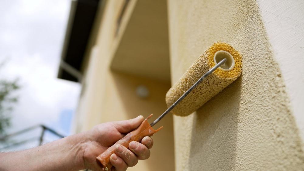 A close-up of a hand using a paint roller to apply light yellow paint to the exterior wall of a building. The focus is on the roller and the textured surface of the wall.