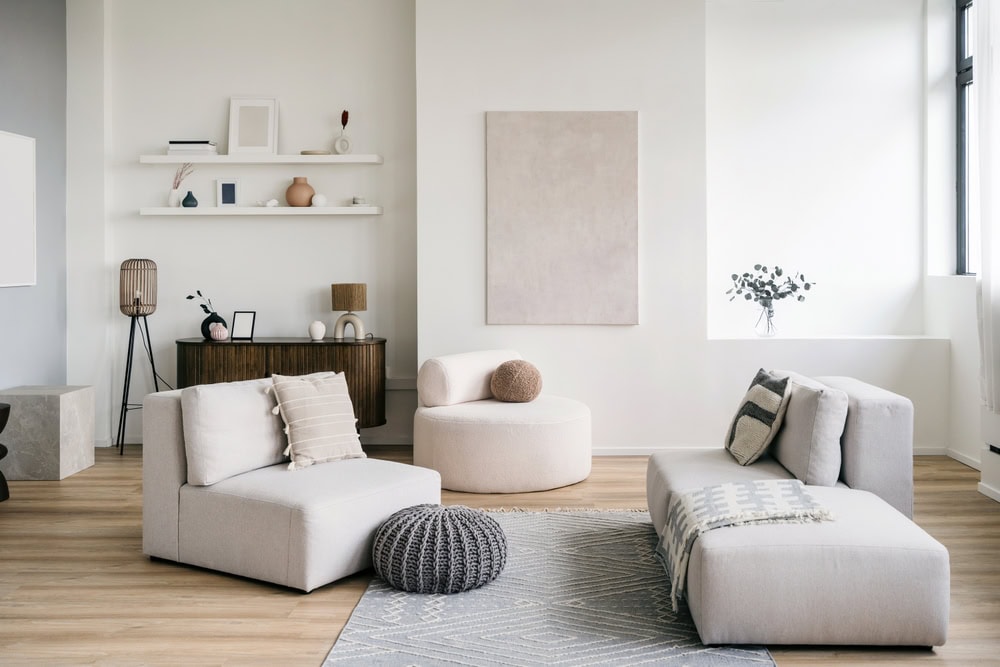 Modern living room with neutral tones, featuring low, light-gray sofas, a round ottoman, a textured pouf, decorative pillows, a patterned rug, wooden cabinet, floating shelves with decor, and a large abstract canvas on the wall.