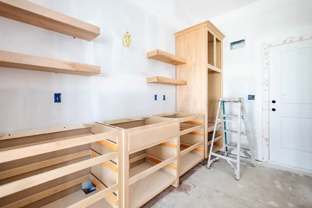 Unfinished kitchen with light wood cabinets and open shelves installed, missing cabinet doors and countertops; a step ladder stands nearby on a bare floor, and unpainted walls with visible outlets await painters in Macomb & Oakland County, MI.