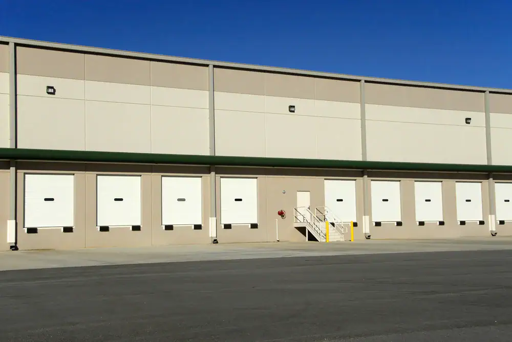 A large beige warehouse building with multiple white loading dock doors, a set of metal stairs leading to a door, and a wide, empty concrete area in front under a clear blue sky—perfect for painters in Macomb & Oakland County, MI.