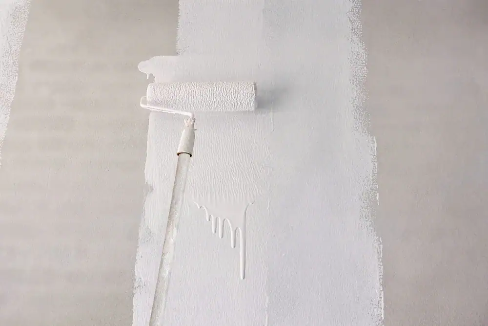 A paint roller is being used to apply white paint on a gray wall by Painters Macomb & Oakland County, MI, with visible paint drips and sections of unpainted surface on either side.